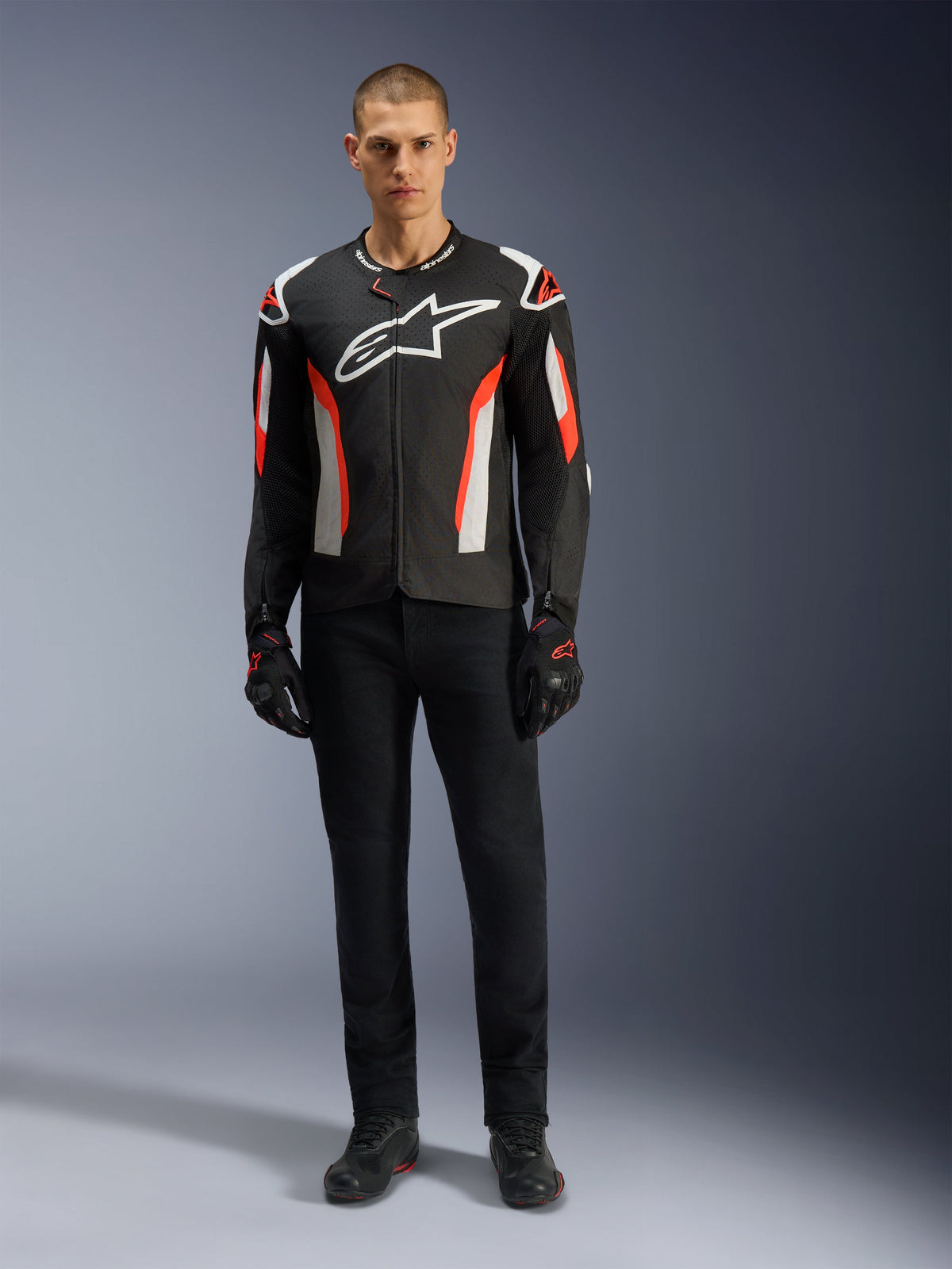 Modèle masculin portant le blouson Alpinestars T-GP Air en Noir Rouge Fluo, blouson de moto en textile avec accents blancs et rouge vif, grand logo Alpinestars blanc sur la poitrine, doté de panneaux de ventilation en mesh, associé à un pantalon technique noir et des gants de moto, équipement de coupe sport