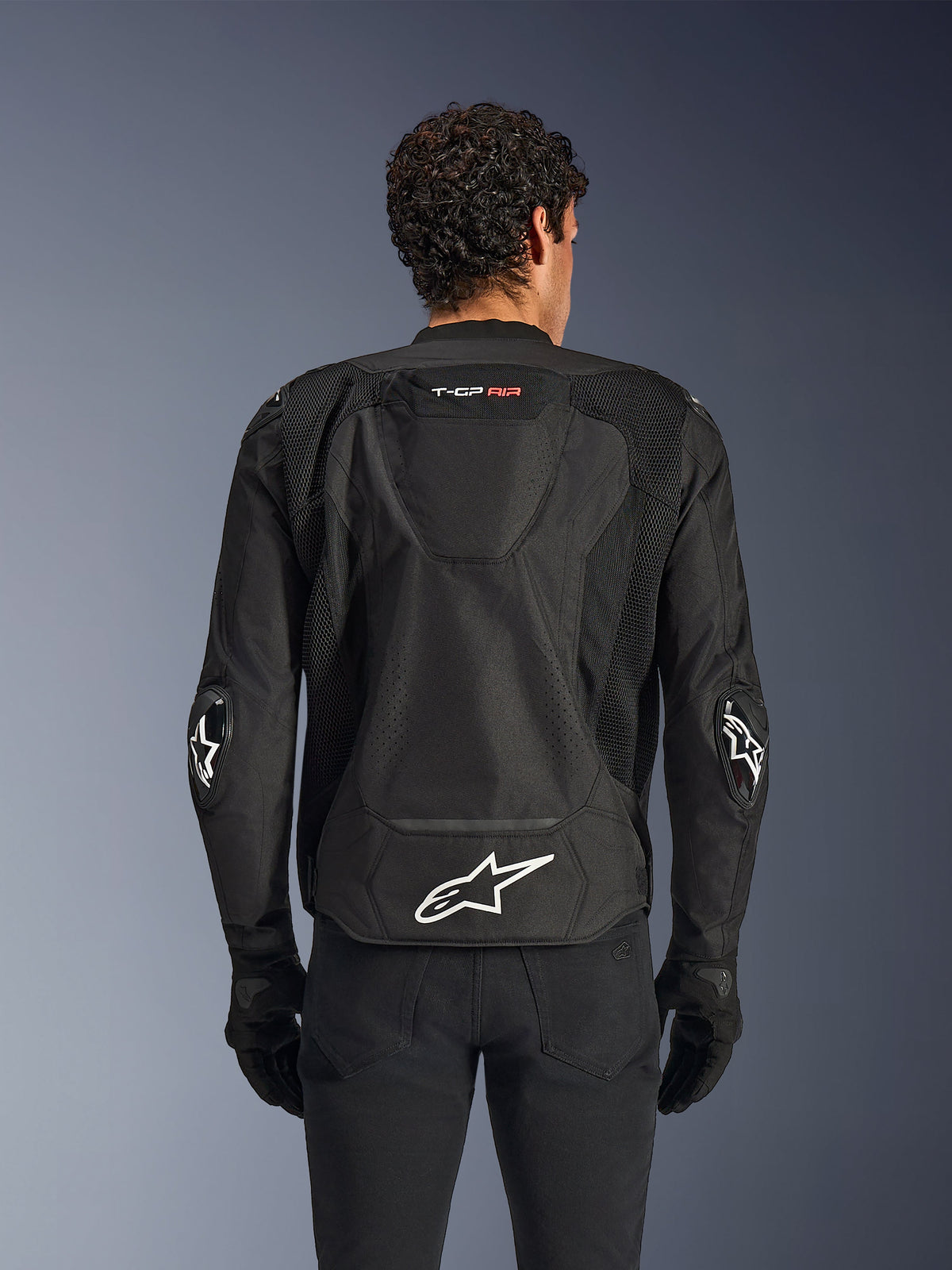 Alpinestars T-GP Air Jacket, noir, vue arrière d'un blouson de moto en textile, doté d'une bosse aérodynamique, de larges panneaux de ventilation en maille et d'un logo Alpinestars blanc dans le bas du dos, porté avec des gants et un pantalon de moto noirs
