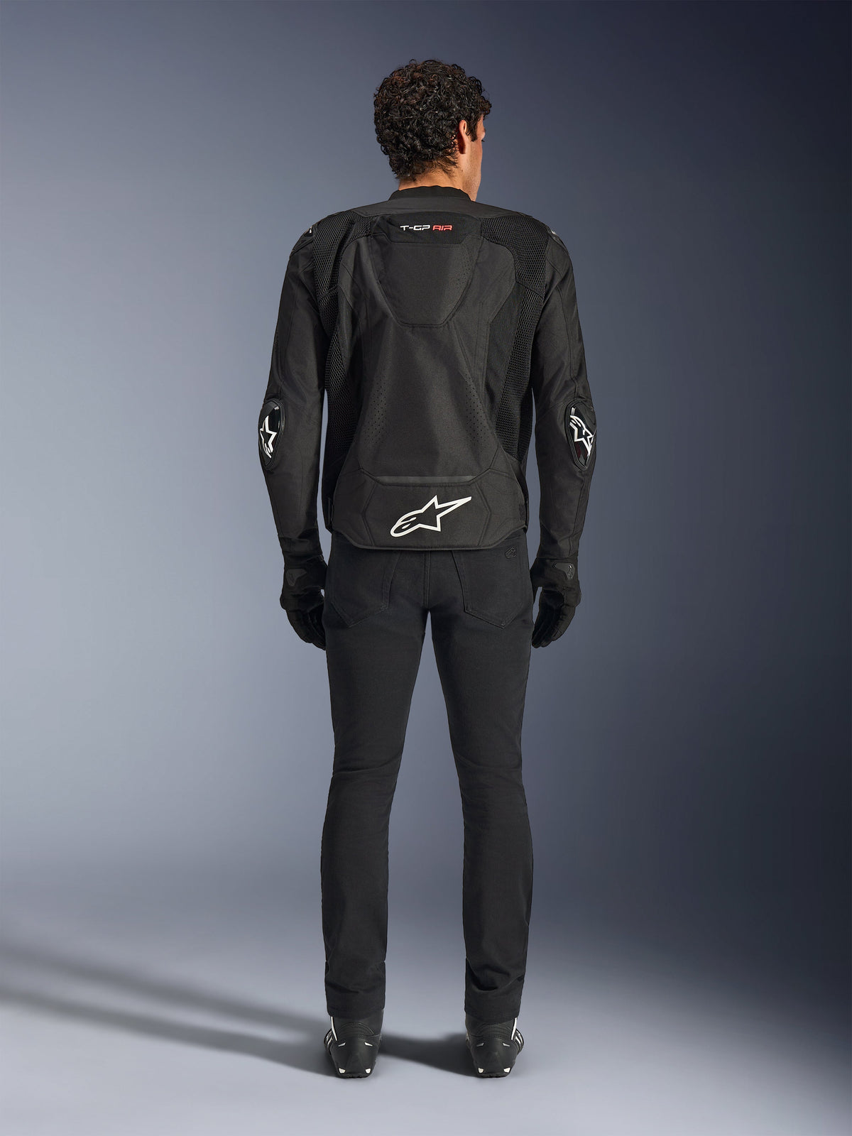 Alpinestars T-GP Air Jacket, noir, vue arrière d'un homme portant la veste de moto textile, présentant des panneaux de ventilation en maille, une bosse aérodynamique et des logos Alpinestars blancs sur le bas du dos et les coudes, portée avec un pantalon et des gants de moto noirs