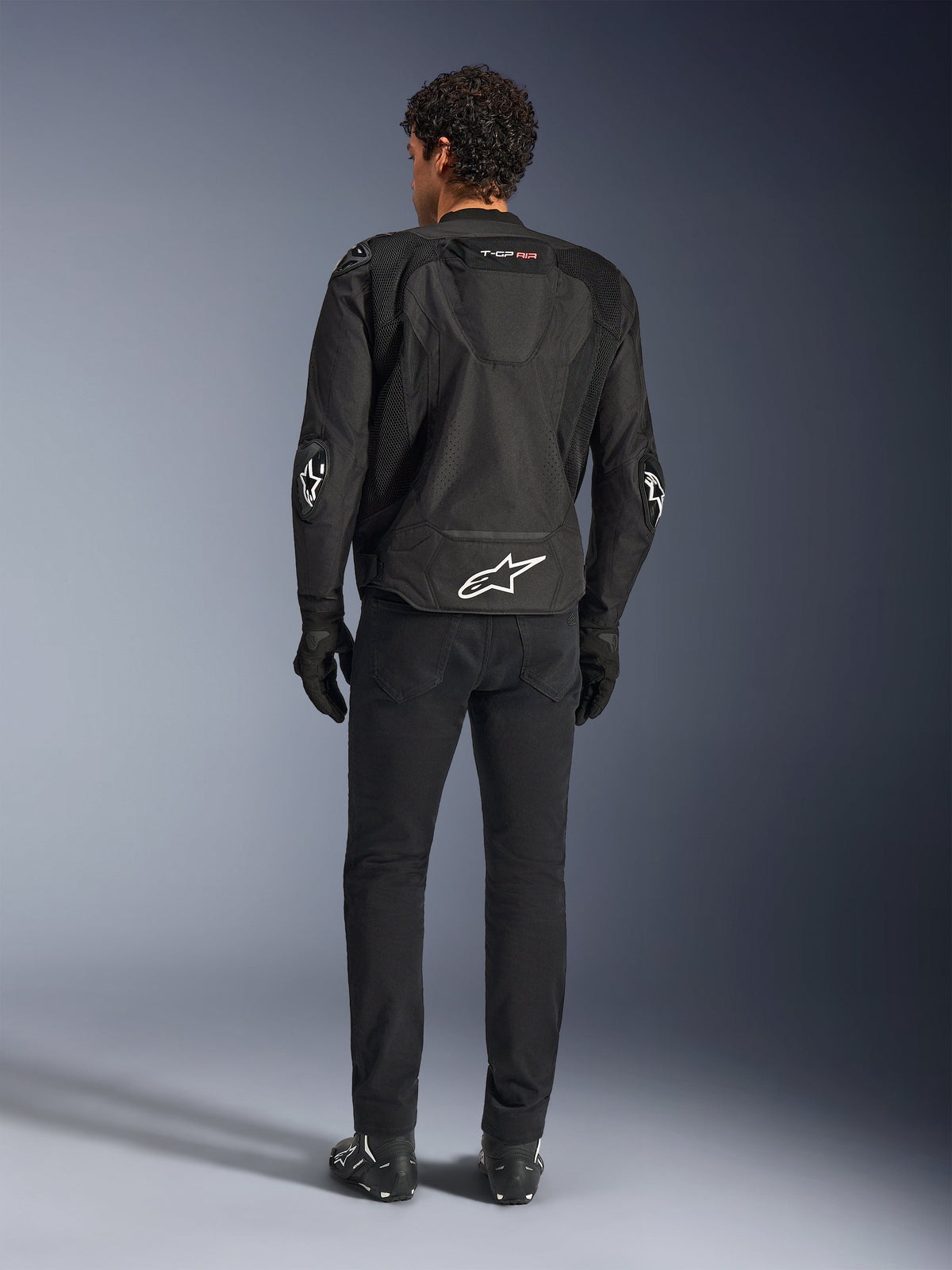 Blouson Alpinestars T-GP Air, noir, vue arrière sur modèle, montrant les panneaux de ventilation en maille 3D et la bosse aérodynamique, avec logo étoile blanc dans le bas du dos et aux coudes, porté avec un pantalon de moto et des gants noirs, équipement de conduite sportive