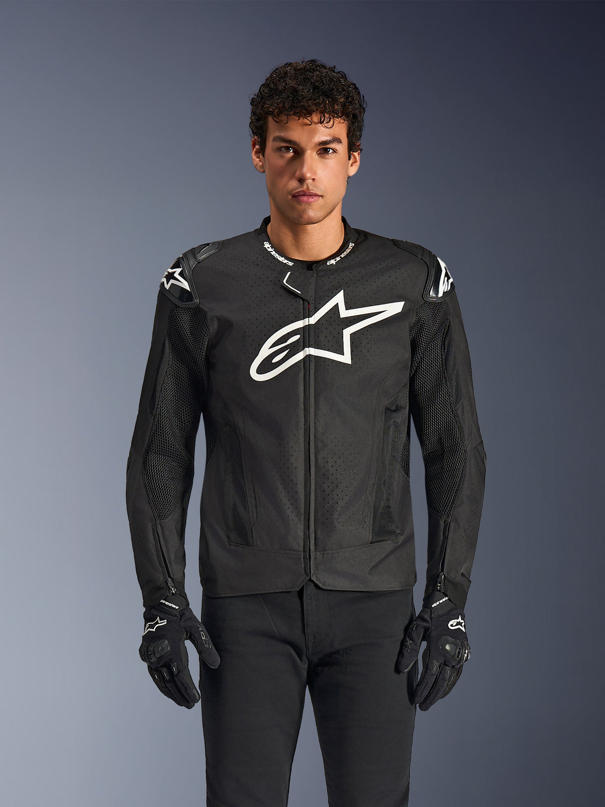 Veste Alpinestars T-GP Air, veste de moto en textile noir avec un grand logo étoile blanc sur la poitrine, dotée de panneaux de ventilation perforés et de mesh 3D, sliders d'épaules externes, portée avec des gants noirs et blancs, design sportif élégant pour la conduite sur route par temps chaud