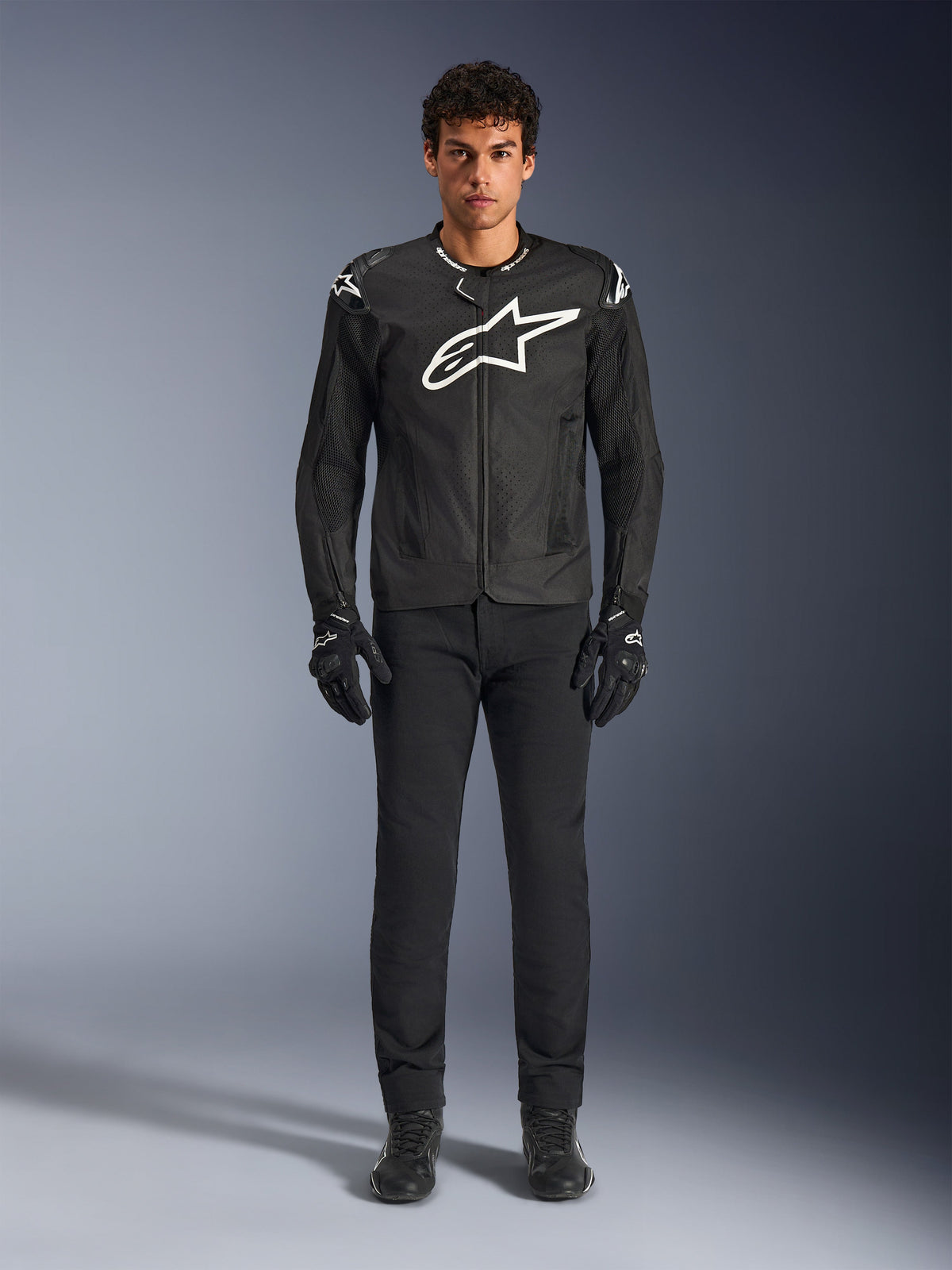 Veste Alpinestars T-GP Air, veste de moto en textile noir avec un grand logo Astars blanc sur la poitrine, portée par un mannequin avec un pantalon et des gants de moto noirs, dotée de panneaux perforés et d'une ventilation en mesh pour la conduite sportive