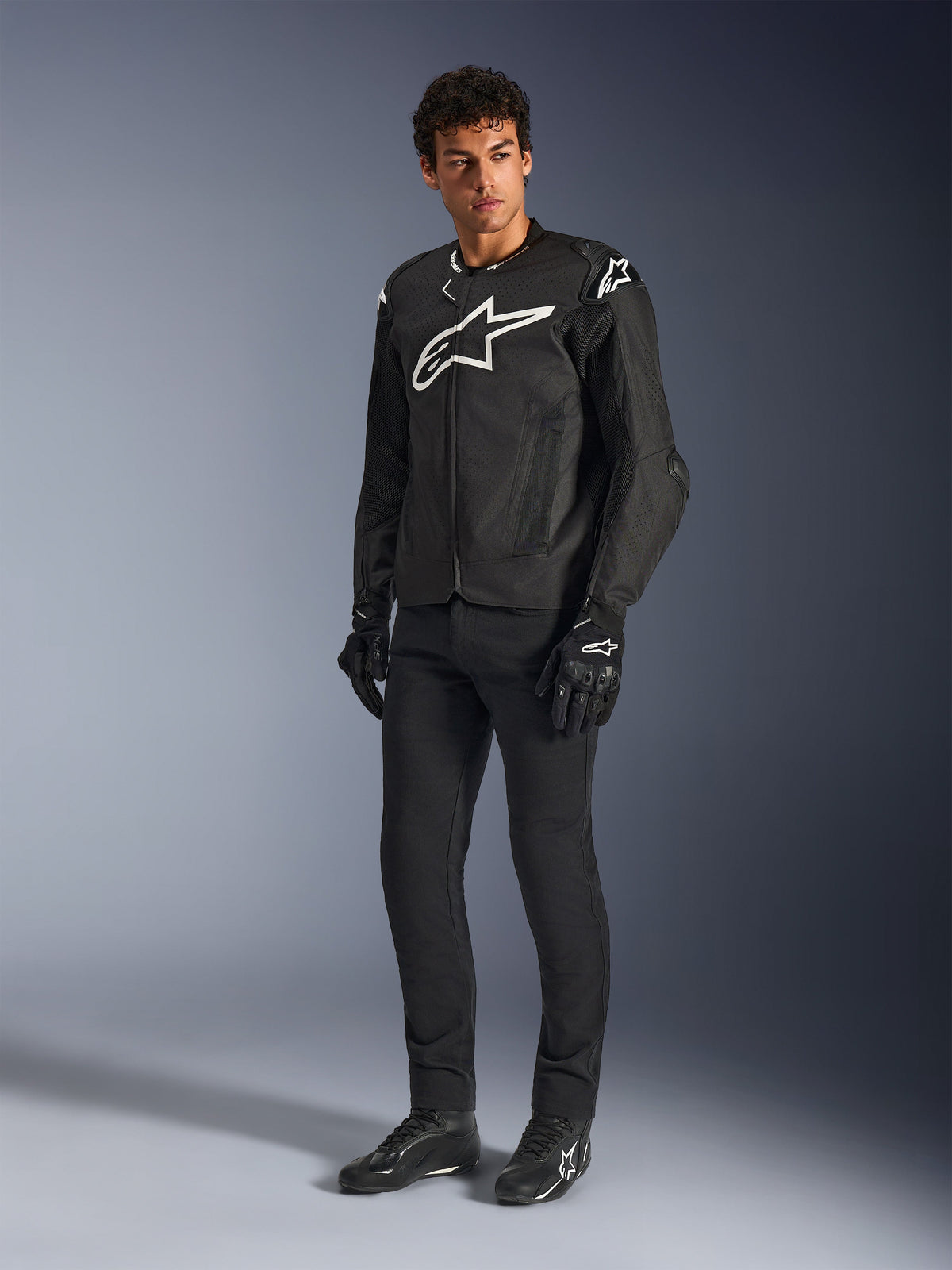 Blouson Alpinestars T-GP Air, veste de moto en textile noir, porté par un mannequin avec pantalon et gants noirs assortis, arborant un logo Alpinestars blanc sur la poitrine et les épaules, des panneaux en maille ventilés et une coupe sportive dérivée de la course