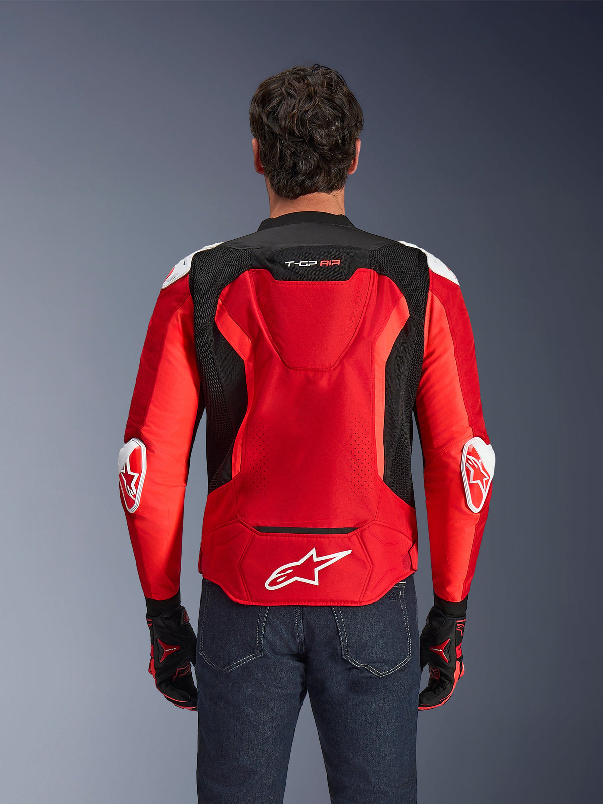 Blouson Alpinestars T-GP Air, veste de moto en textile, rouge vif et noir avec des accents blancs, vue de dos montrant la bosse aérodynamique et les panneaux de ventilation en maille, arborant le logo étoile Alpinestars et le marquage T-GP Air, porté avec un jean foncé et des gants de course.