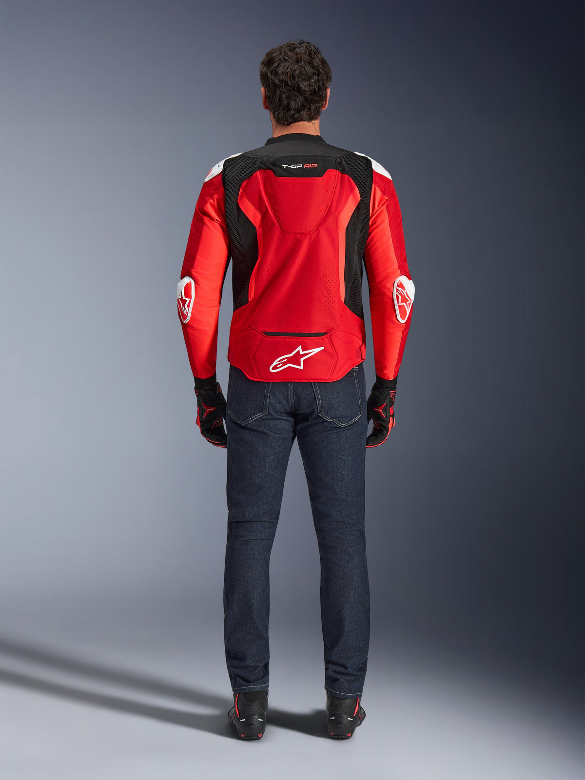 Veste Alpinestars T-GP Air, vue arrière, rouge vif et noir, avec logo blanc dans le bas du dos et marquage « T-GP AIR », portée avec un jean en denim bleu foncé et des gants de moto noirs et rouges, équipement de moto de sport haute performance