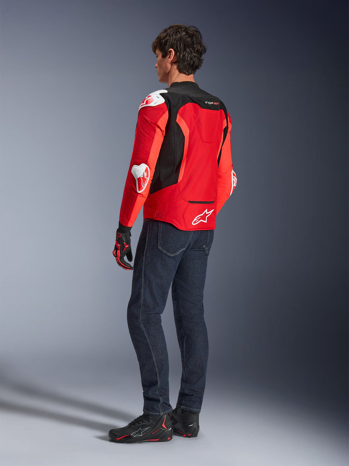 Veste Alpinestars T-GP Air, vue arrière d'un homme portant une veste textile rouge vif et rouge fluo avec des panneaux en maille noire et des sliders de protection blancs, associée à un jean bleu foncé et des chaussures de moto noires, équipement de moto sportive