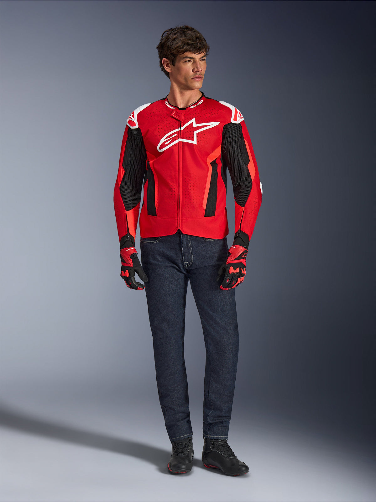 Veste Alpinestars T-GP Air, rouge vif, rouge fluo et noir, modèle masculin portant une tenue de moto complète avec jean bleu foncé et gants assortis, veste textile sportive avec panneaux en maille perforée et logo Astars blanc, photo de studio.