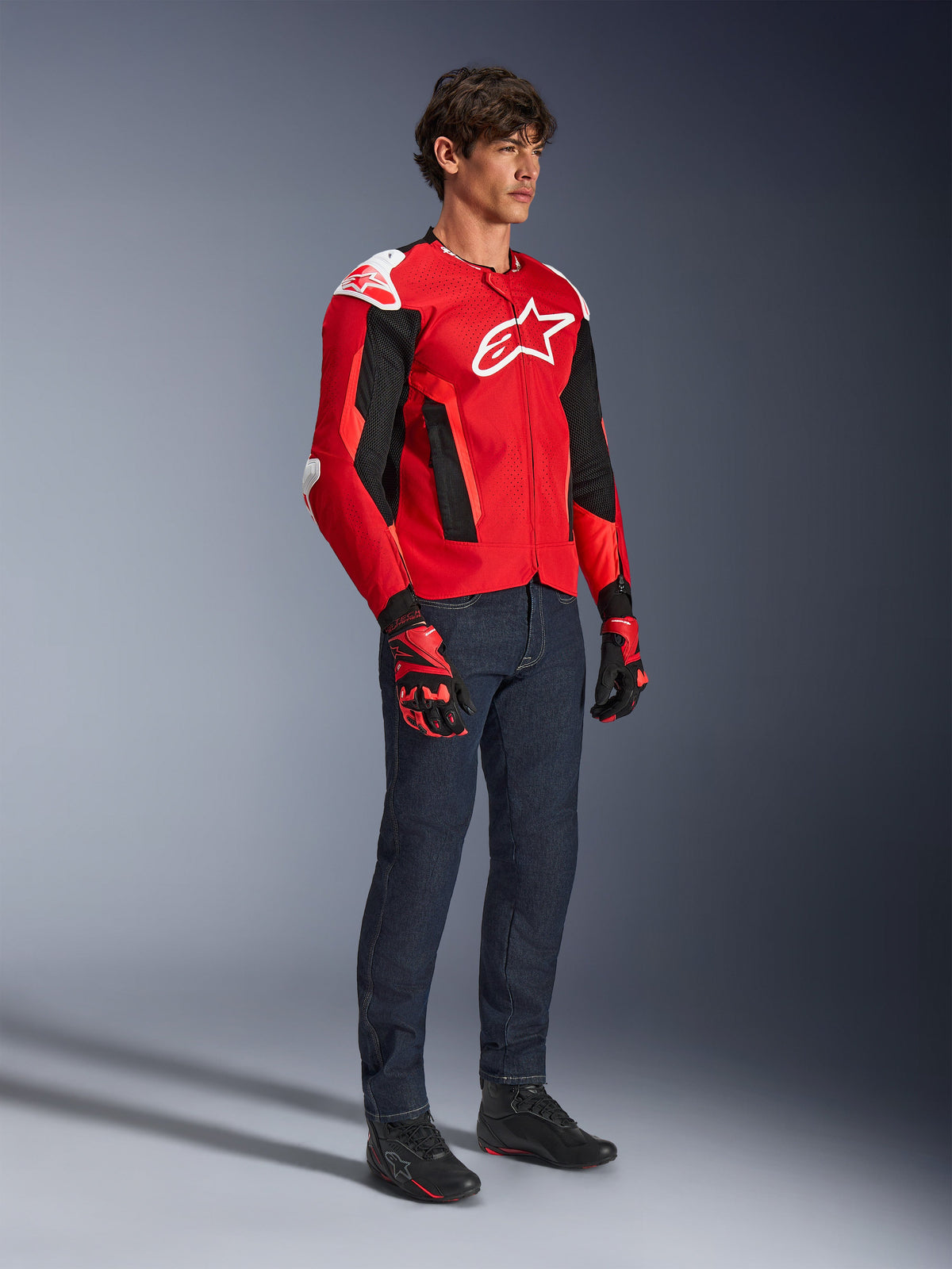 Veste Alpinestars T-GP Air, veste de moto sport, rouge vif et noir avec des accents blancs, portée par un mannequin avec un jean bleu foncé et des gants de course rouges, dotée de panneaux en maille à haut flux et de sliders d'épaule blancs, coupe sport aérodynamique pour la conduite par temps chaud.
