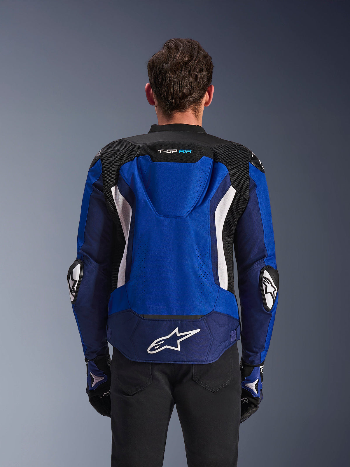 Blouson Alpinestars T-GP Air, blouson de moto sport en textile, bleu éclatant, marine et blanc, vue arrière présentant la bosse aérodynamique intégrée et le grand logo blanc sur le bas du dos, porté avec des gants de moto assortis bleus et noirs