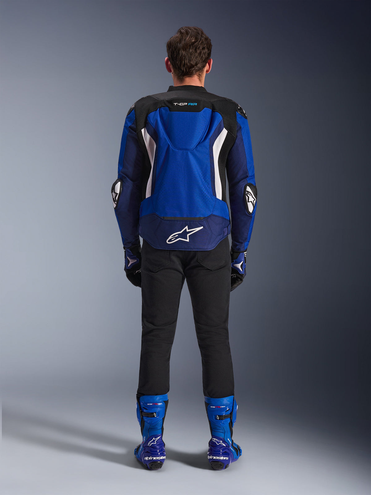 Blouson Alpinestars T-GP Air, Bleu Brillant Marine Blanc, vue de dos montrant la bosse aérodynamique et les panneaux de ventilation en maille, logo Alpinestars blanc en bas du dos, porté avec un pantalon noir et des bottes de course bleues