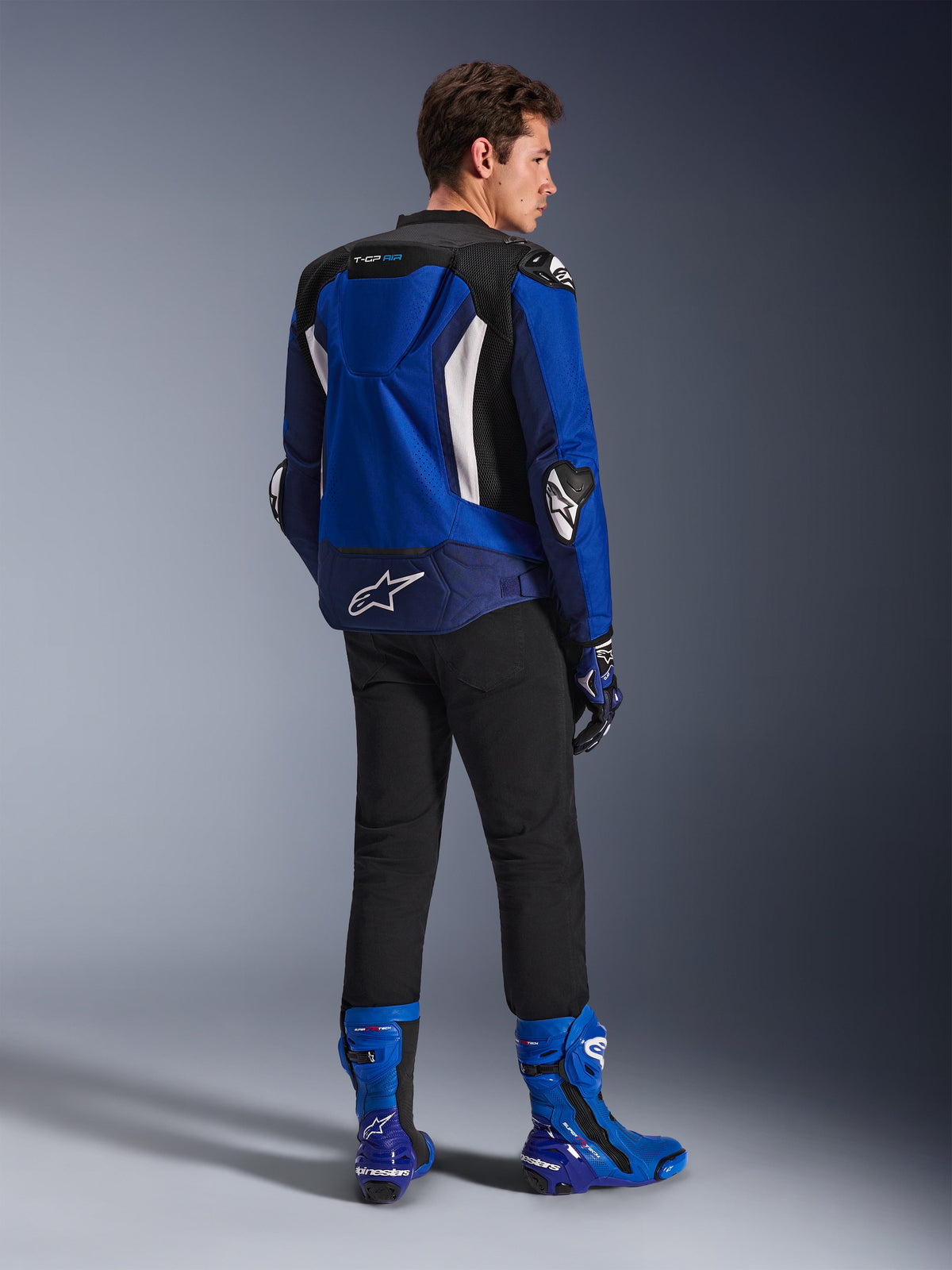 Veste Alpinestars T-GP Air, veste textile sport, bleu vif, bleu marine et blanc, vue de dos sur un mannequin portant un pantalon noir et des bottes bleues et blanches assorties, avec logo Alpinestars blanc sur le bas du dos, panneaux de ventilation en mesh et bosse aérodynamique