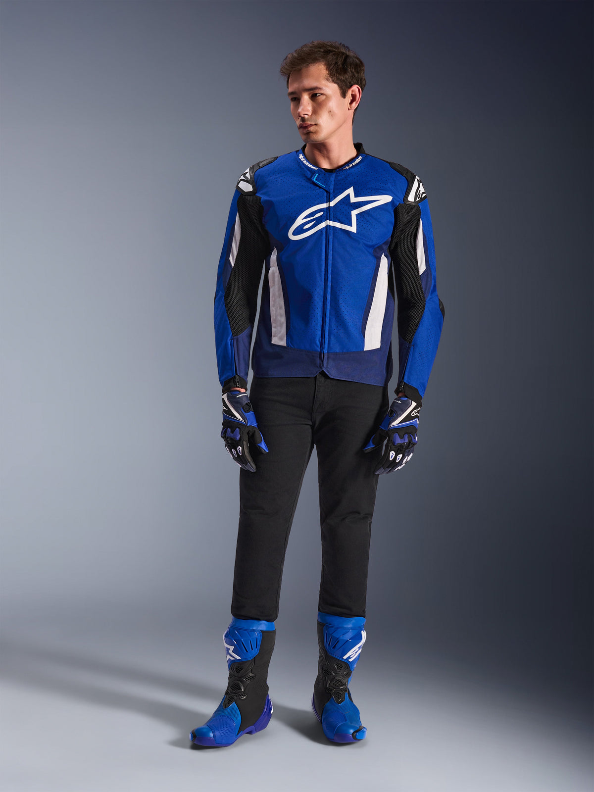 Modèle masculin portant la veste Alpinestars T-GP Air en bleu vif, bleu marine et blanc, associée à un pantalon noir, des gants de course assortis et des bottes de moto bleues et noires, photo de studio présentant une tenue de route sportive complète.