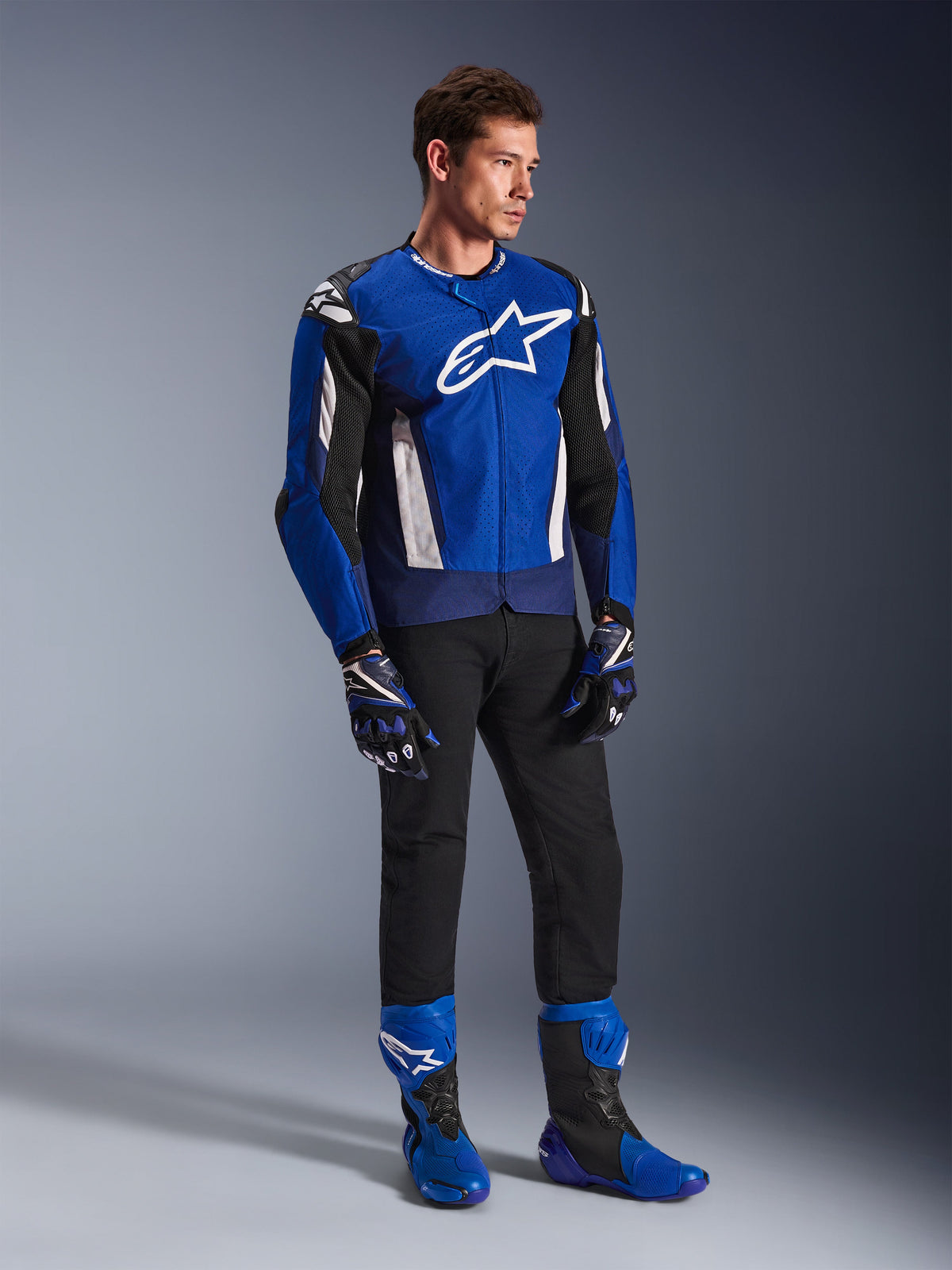 Veste Alpinestars T-GP Air, veste de moto sport en textile, bleu vif, bleu marine et blanc, portée avec des gants bleus et noirs assortis, un pantalon noir et des bottes de course bleues, avec logo blanc sur la poitrine et panneaux en maille technique, photo de studio en pied