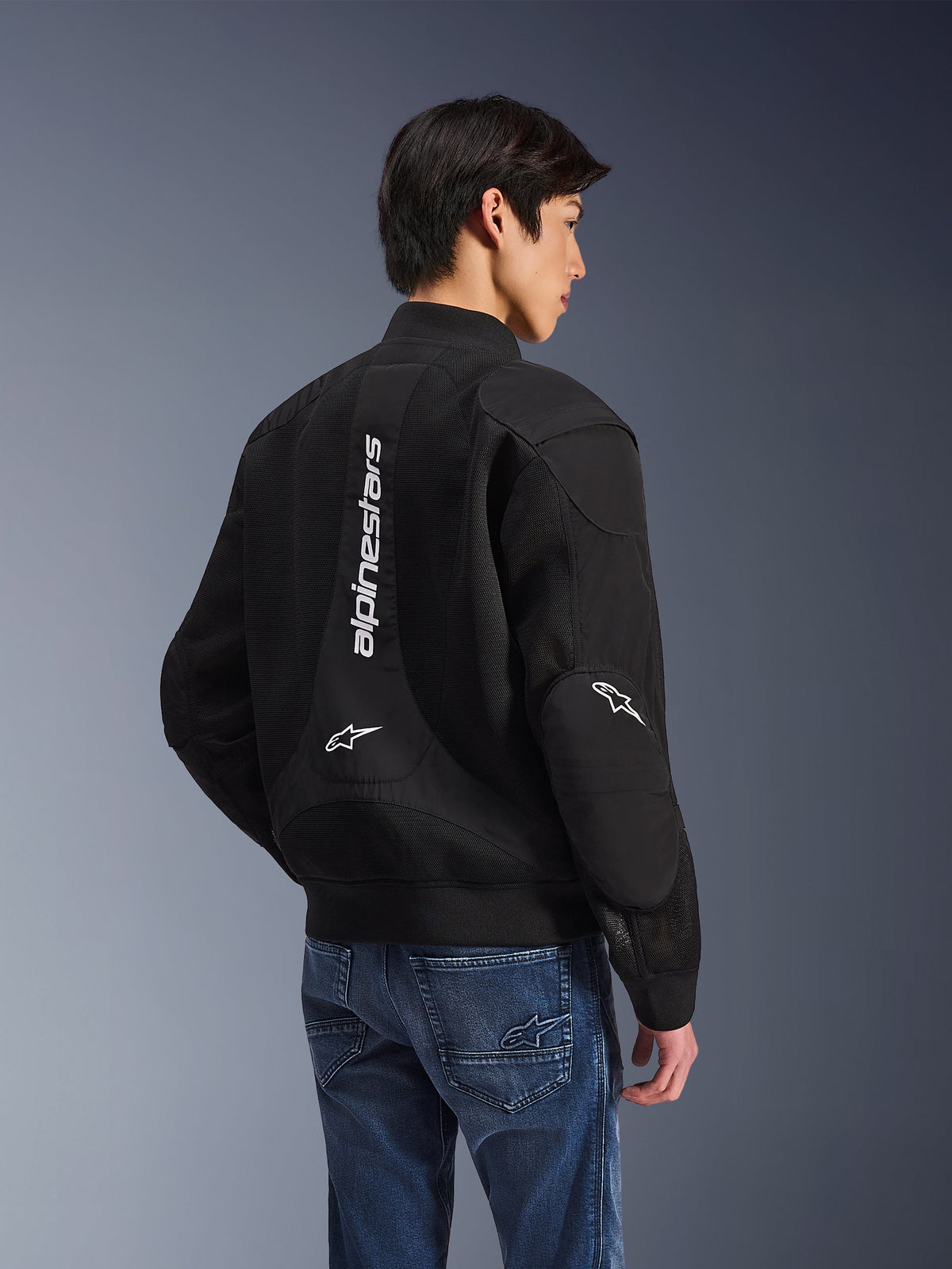 Alpinestars Flight Air Jacket, veste de moto noire, vue arrière montrant un grand logo blanc vertical et des panneaux de ventilation en maille, style bomber avec ourlet côtelé, portée avec un jean bleu, équipement de conduite urbain contemporain