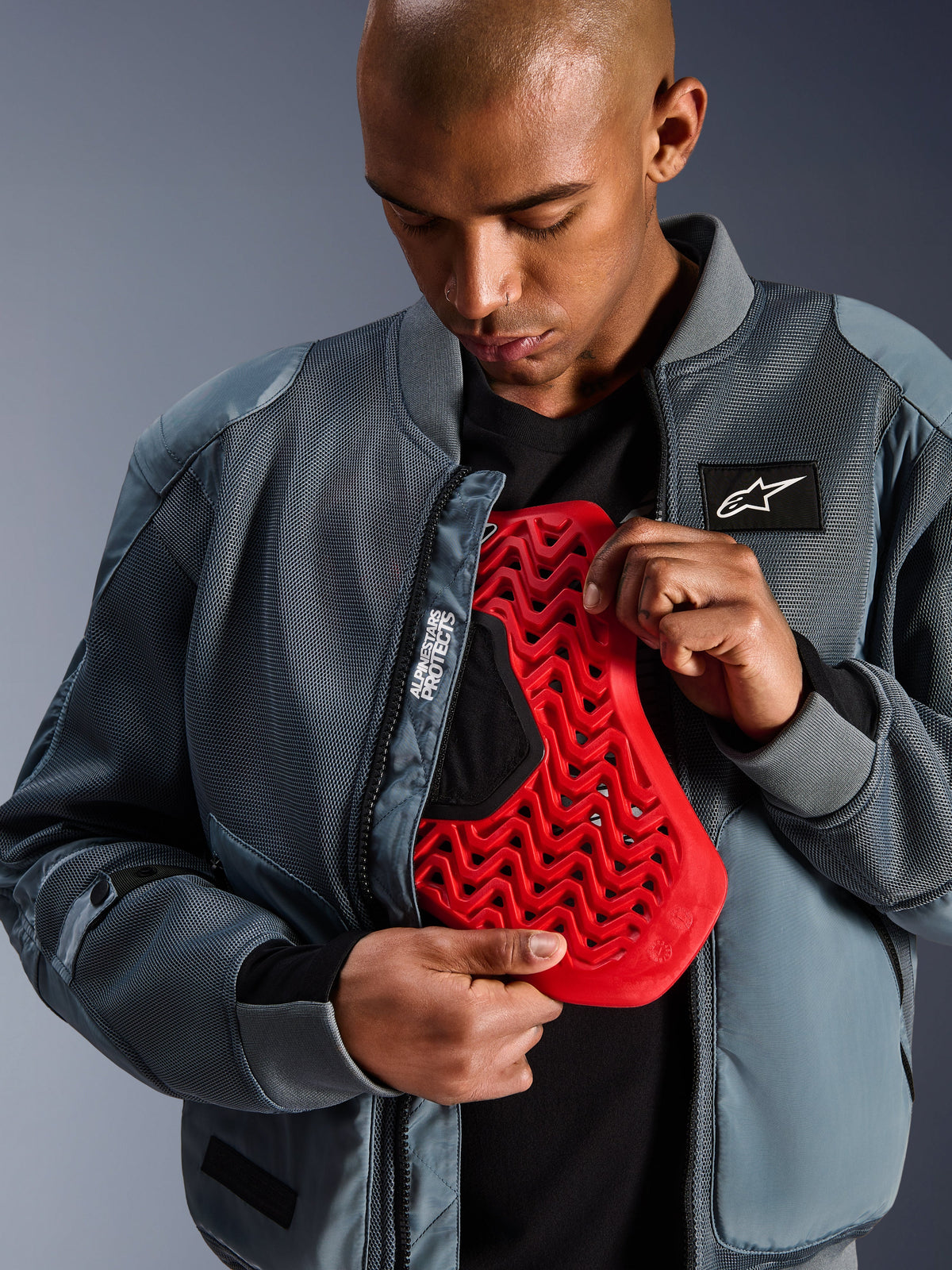 Veste Alpinestars Flight Air en Storm Gray, veste bomber de moto urbaine avec panneaux en maille 3D pour la ventilation, ouverte pour révéler une protection dorsale Nucleon rouge, arborant le marquage « Alpinestars Protects » et une esthétique streetwear moderne