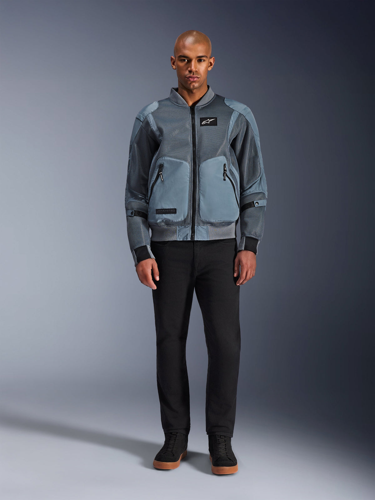 Veste Alpinestars Flight Air en Storm Gray, veste de moto urbaine d'inspiration bomber dotée de panneaux en maille 3D et de poches avant, portée par un homme avec un pantalon noir et des baskets montantes noires, vêtement de conduite moderne au style urbain