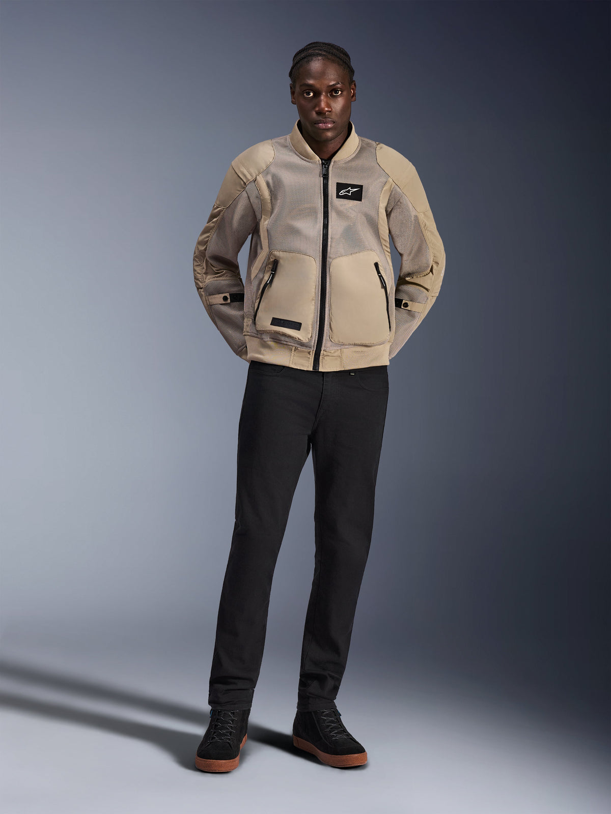 Model wearing the Alpinestars Flight Air Jacket in Brown, urban motorcycle jacket with extended 3D mesh panels, bomber-inspired ribbed collar and cuffs, styled with black pants and black riding shoes, contemporary street-ready gear