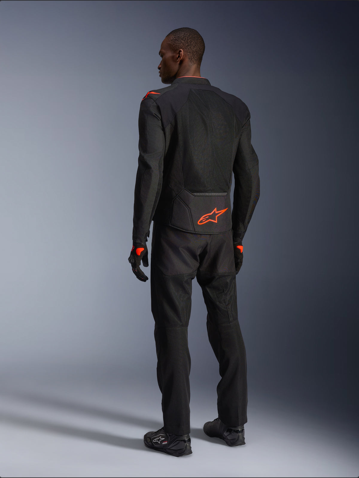 Pilote homme vu de dos portant la veste Alpinestars Viper Air V4, noir et rouge fluo, logo Astars orange sur le bas du dos et les épaules, construction en textile mesh, avec pantalon et bottes de moto noirs assortis