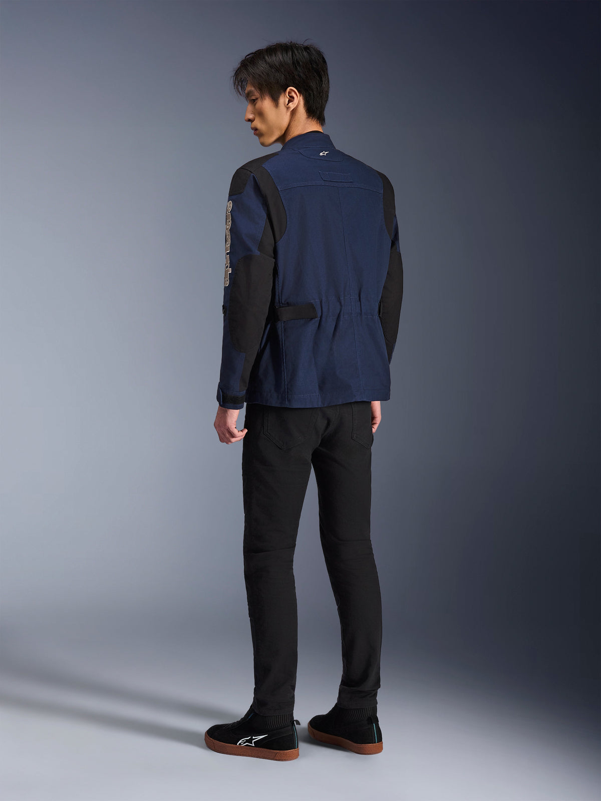 Male model viewed from the back wearing the Alpinestars Dice Jacket in Dark Blue and Black, paired with black pants and black sneakers, urban motorcycle field jacket featuring box-pleated shoulders and adjustable waist tabs.