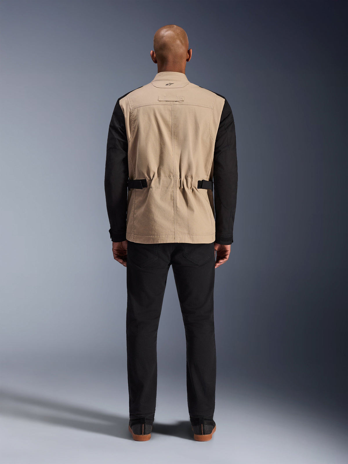 Veste Dice Alpinestars, veste de moto urbaine, beige et noir, vue de dos sur mannequin, dotée d'une construction en sergé de polycoton, de sangles de réglage à la taille et d'un petit logo brodé sur le col, portée avec un pantalon noir