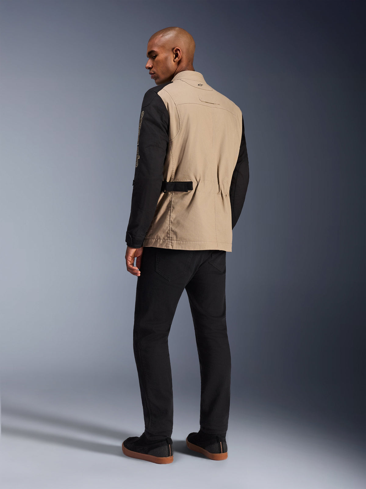Veste Alpinestars Dice, veste de moto urbaine, Aluminum Black avec buste beige et manches noires, vue de dos sur modèle, design robuste d'inspiration workwear avec ajustements à la taille, associée à un pantalon de moto noir.