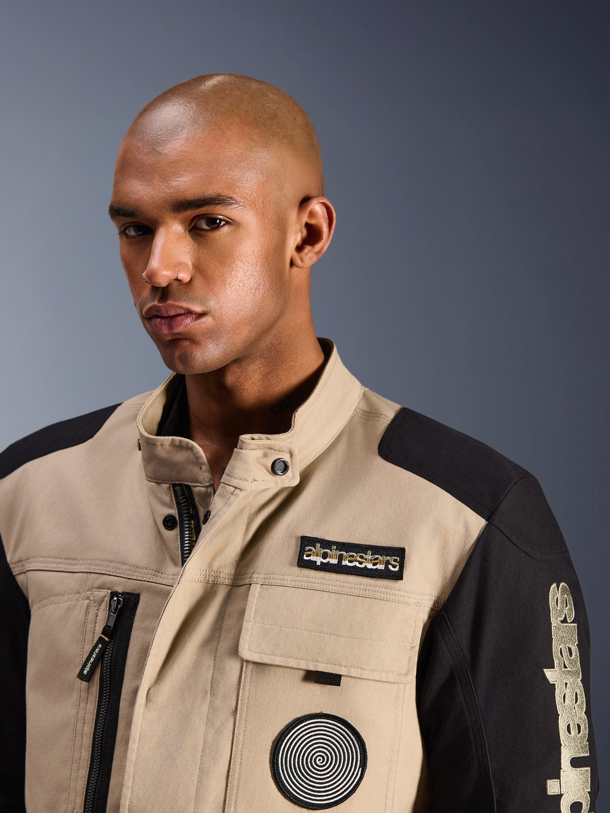 Mannequin homme portant la veste Alpinestars Dice en Aluminum Black, veste de moto urbaine avec panneaux contrastés beige et noir, dotée d'un écusson Alpinestars brodé sur la poitrine et d'un logo spirale, design robuste inspiré du workwear avec un col haut.