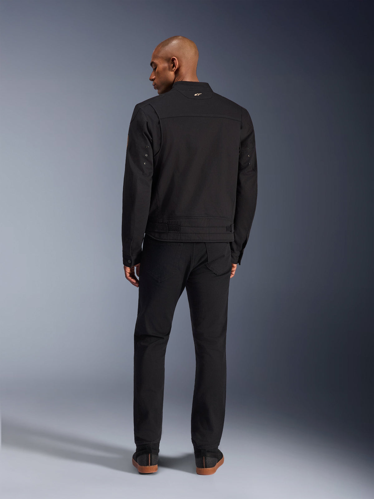Alpinestars Bruiser Jacket in Black, back view of male model wearing the urban motorcycle jacket with matching black pants, featuring a small gold logo on the collar and adjustable waist straps, workwear-inspired aesthetic