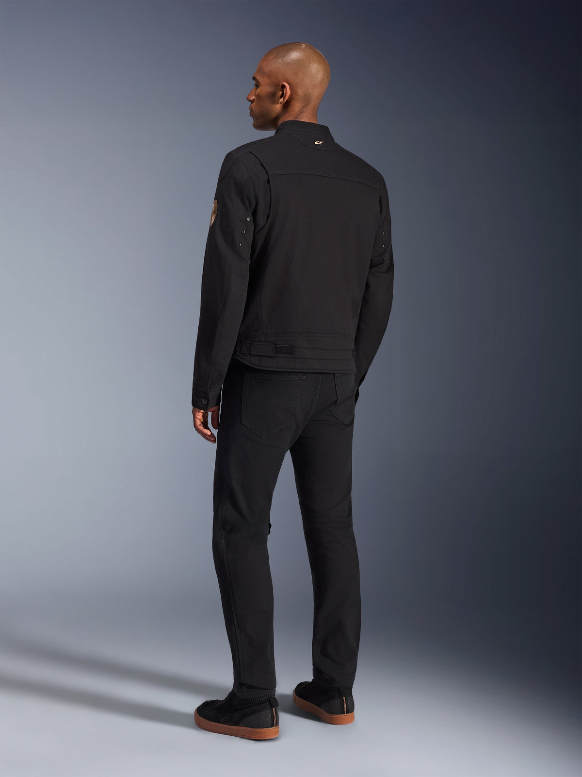 Alpinestars Bruiser Jacket, Urban Black Motorcycle Jacket, back view on a model, classic workwear styling, featuring pleated shoulder panels and Oscar spiral patch on the sleeve, worn with black pants and casual black shoes with gum soles.