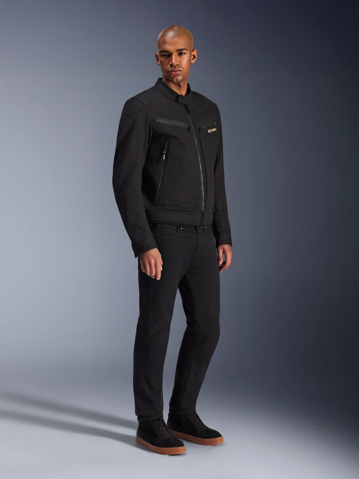 Alpinestars Bruiser Jacket, Urban Motorcycle Jacket, Black, worn by a model with black pants and sneakers, featuring zippered chest pockets, polycotton twill fabric, and a sleek workwear-inspired design for urban riding