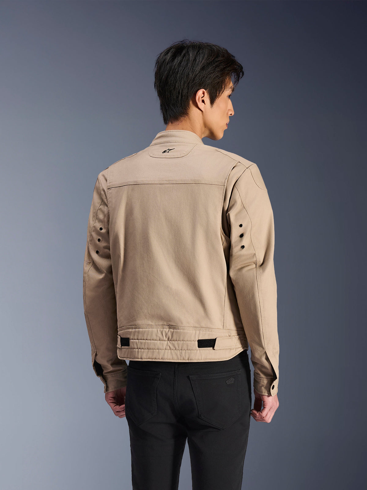 Veste Alpinestars Bruiser, veste de moto urbaine marron, vue arrière montrant les épaules plissées articulées et les sangles de réglage à la taille, portée avec un pantalon noir, équipement de moto contemporain inspiré du style workwear