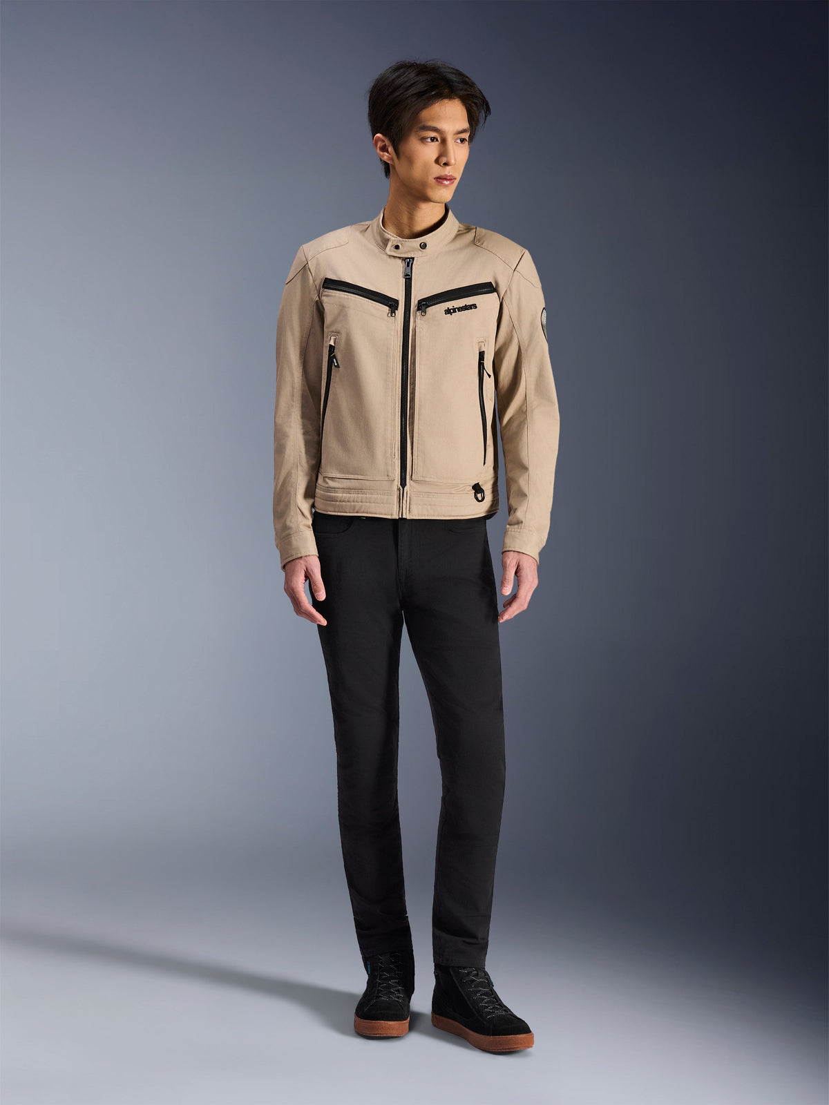 Veste Alpinestars Bruiser, veste de moto urbaine en Aluminum, portée avec un pantalon noir et des baskets noires, dotée de poches zippées noires et d'un col à bouton-pression, design contemporain inspiré du vêtement de travail pour la conduite urbaine
