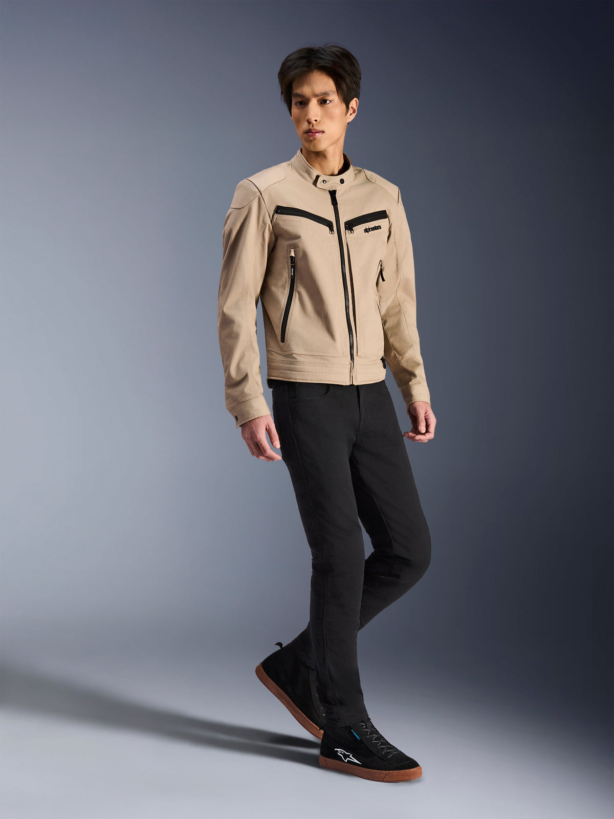 Veste Alpinestars Bruiser, veste de moto urbaine, beige, portée par un mannequin homme avec un pantalon slim noir et des baskets noires, construction textile inspirée du vêtement de travail avec des zips noirs contrastés, style urbain contemporain.