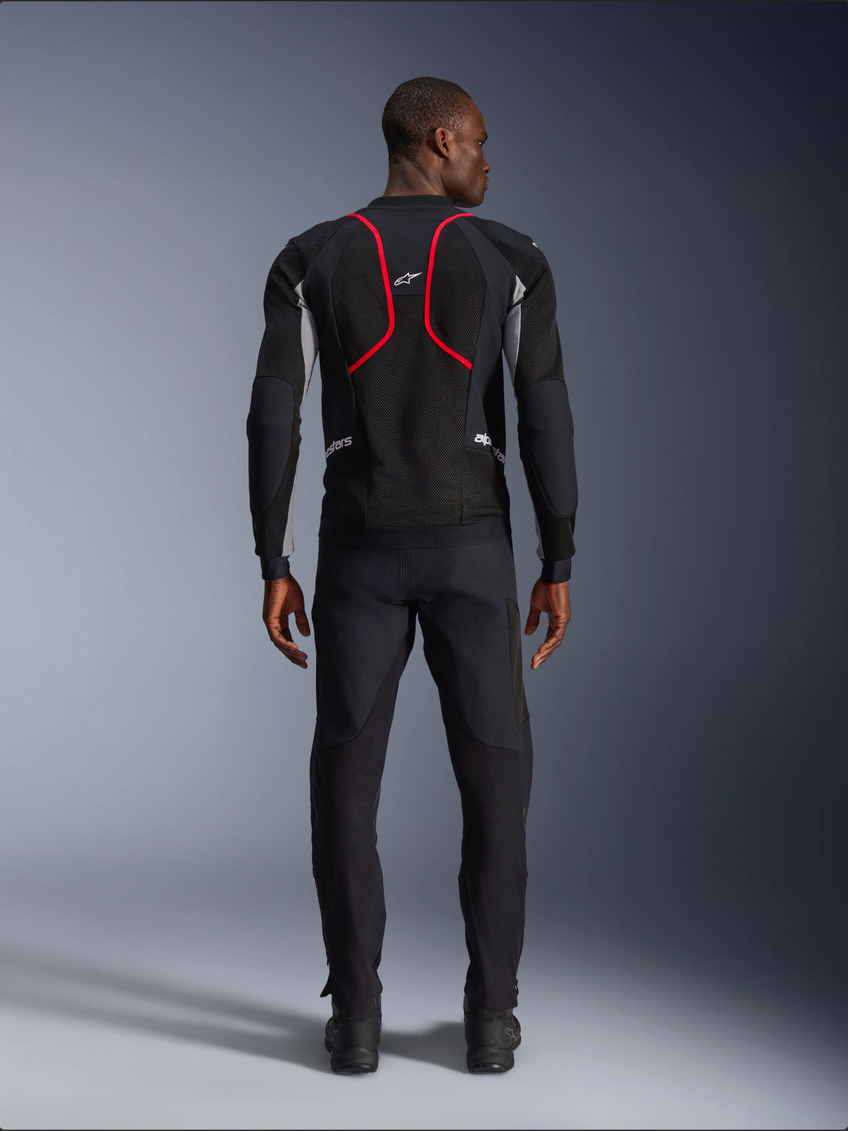 Modèle vu de dos portant la veste Alpinestars Troop-Air Pro, noire avec des accents gris cendré et rouge vif, dotée d'une construction en maille technique avec un liseré rouge proéminent dans le dos, logo Alpinestars sur le haut du dos, associée à un pantalon de moto noir, veste de protection aérée premium pour la conduite sur route