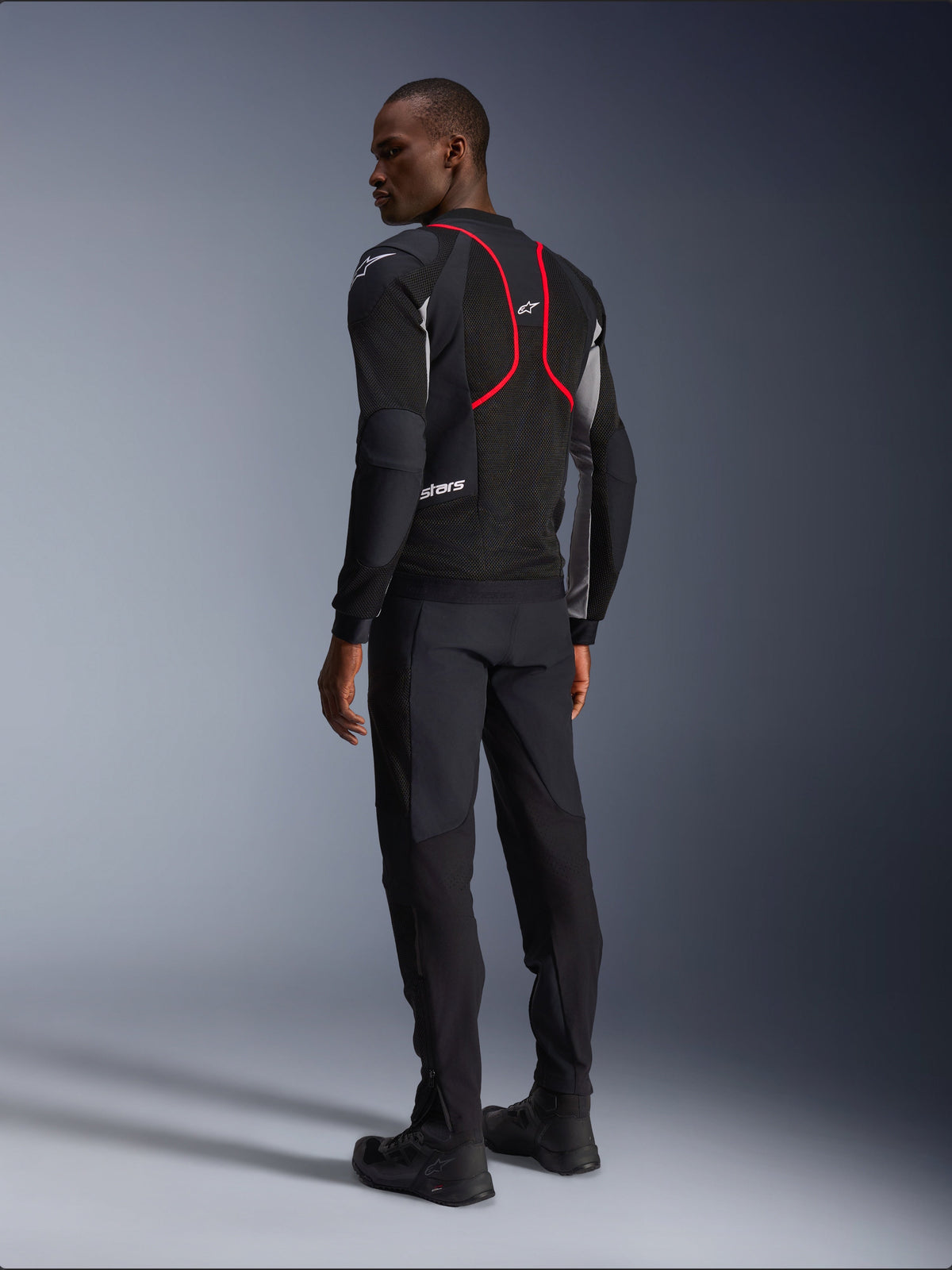 Veste Alpinestars Troop-Air Pro, veste de moto, noir, gris cendré et rouge vif, vue de dos sur mannequin, avec lignes d'accent rouges et panneaux de ventilation en mesh, portée avec un pantalon et des bottes noirs, style moto urbain