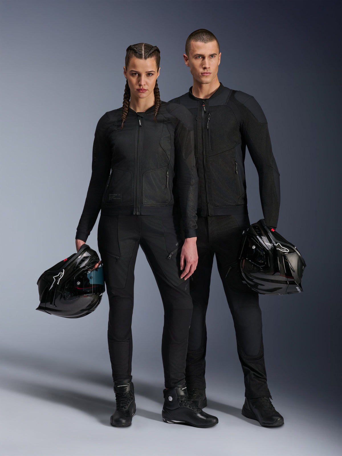 Male and female models wearing Alpinestars Troop-Air Pro Jackets in black, technical mesh armored riding gear paired with matching black pants and boots, each holding a black helmet, showcasing coordinated urban motorcycle apparel.