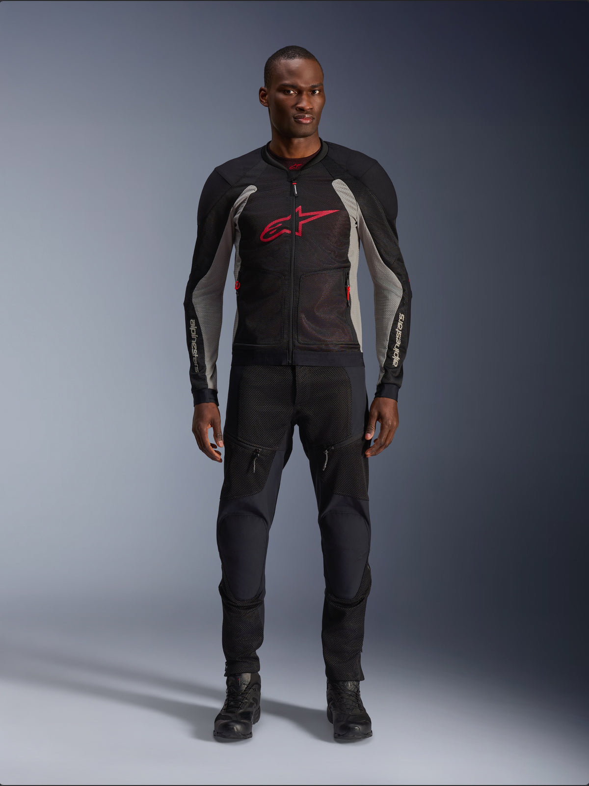 Mannequin homme portant la veste Alpinestars Troop-Air en Noir Gris Cendré et Rouge Vif, veste de moto technique en maille présentant un logo rouge proéminent sur la poitrine, assortie d'un pantalon et de bottes de moto noirs, tenue de route urbaine