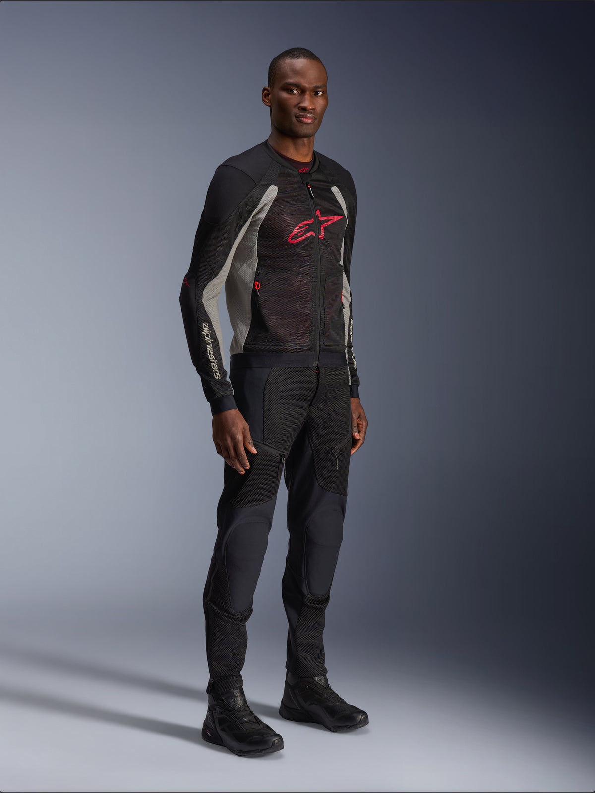 Veste Alpinestars Troop-Air, veste de moto en maille technique noir, gris cendré et rouge vif, portée par un mannequin avec pantalon et bottes noirs assortis, arborant un logo Alpinestars rouge sur la poitrine, équipement de conduite urbain contemporain