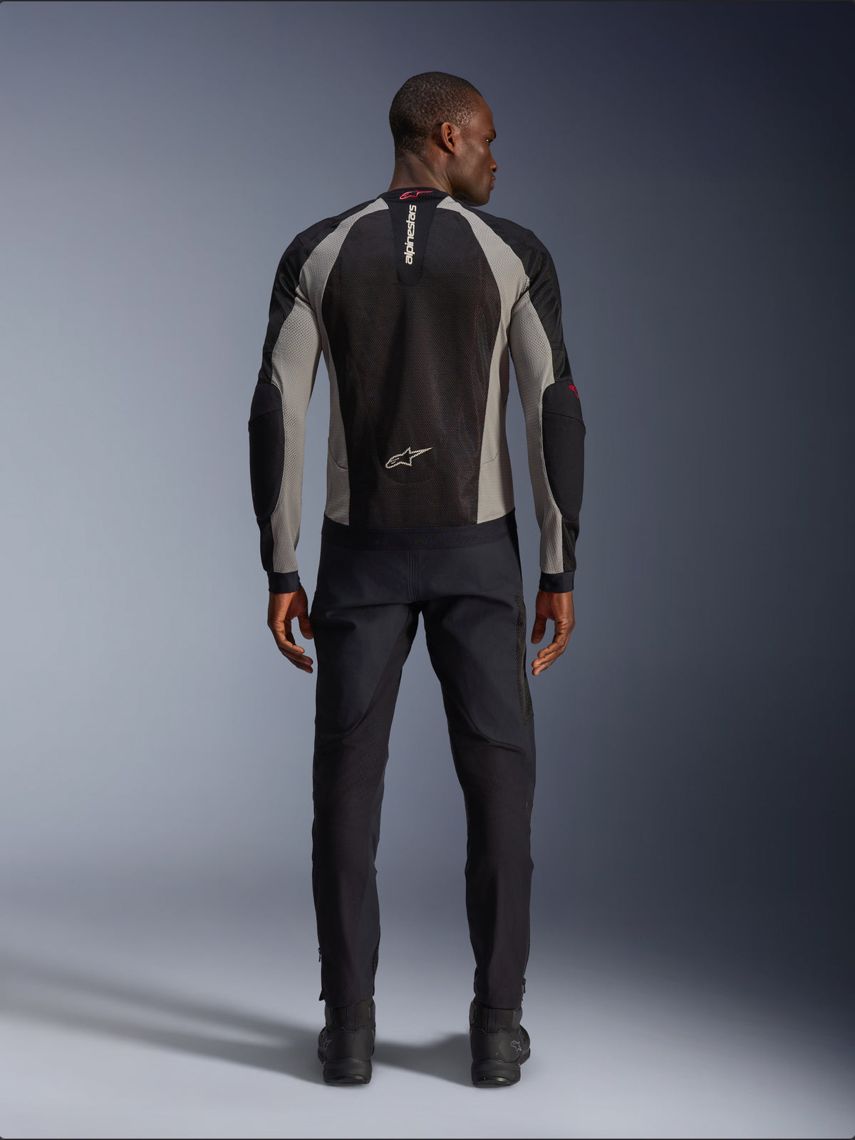 Veste Alpinestars Troop-Air, veste de moto noir, gris cendré et rouge vif, vue arrière sur un modèle masculin, construction en maille légère pour la ventilation, logos Alpinestars sur le col et le bas du dos, portée avec un pantalon de moto noir
