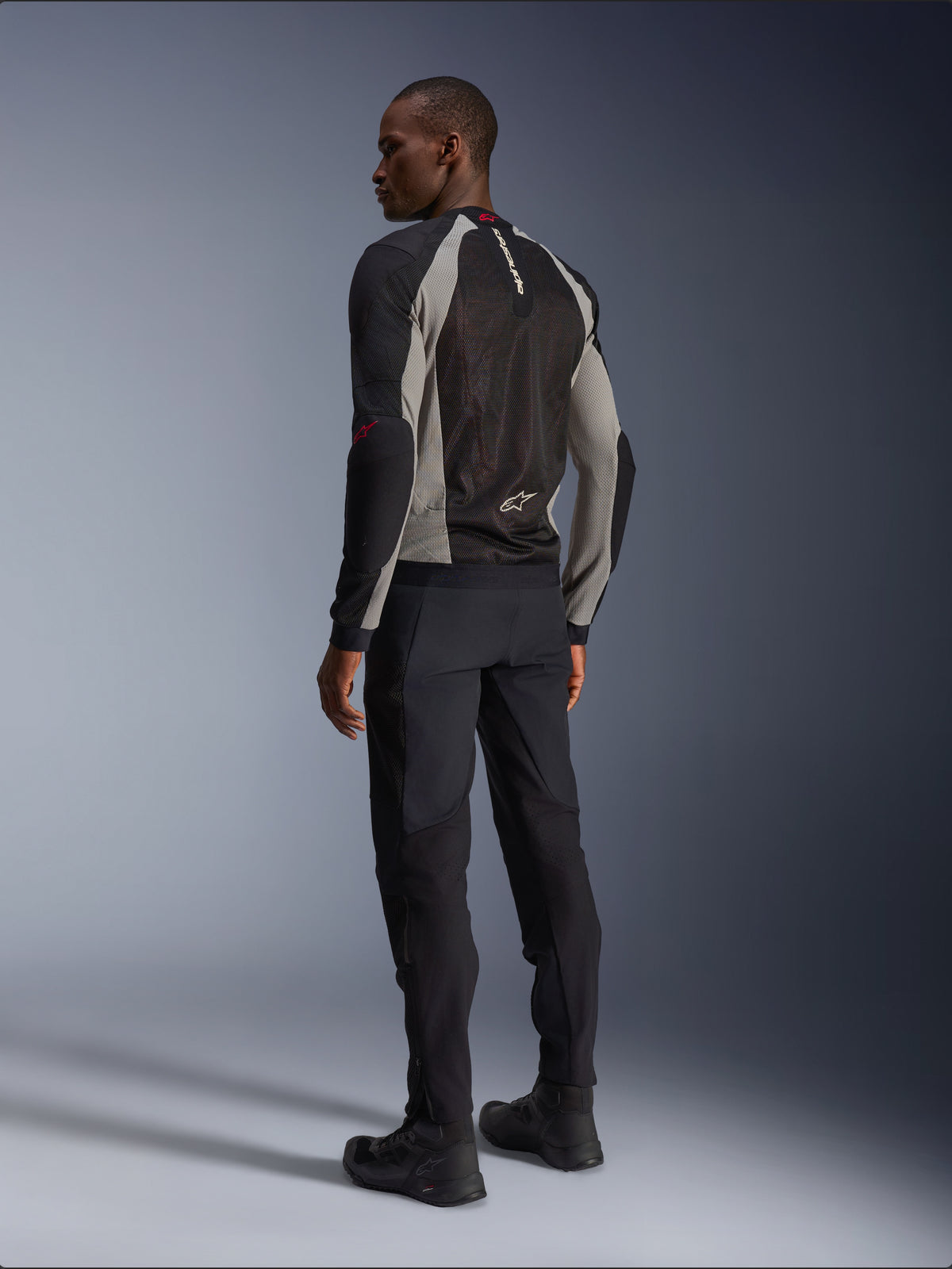 Veste Alpinestars Troop-Air vue de dos, coloris noir, gris cendre et rouge vif, construction technique en maille pour la ventilation, associée à un pantalon de moto noir et des bottes de pilotage, style urbain contemporain