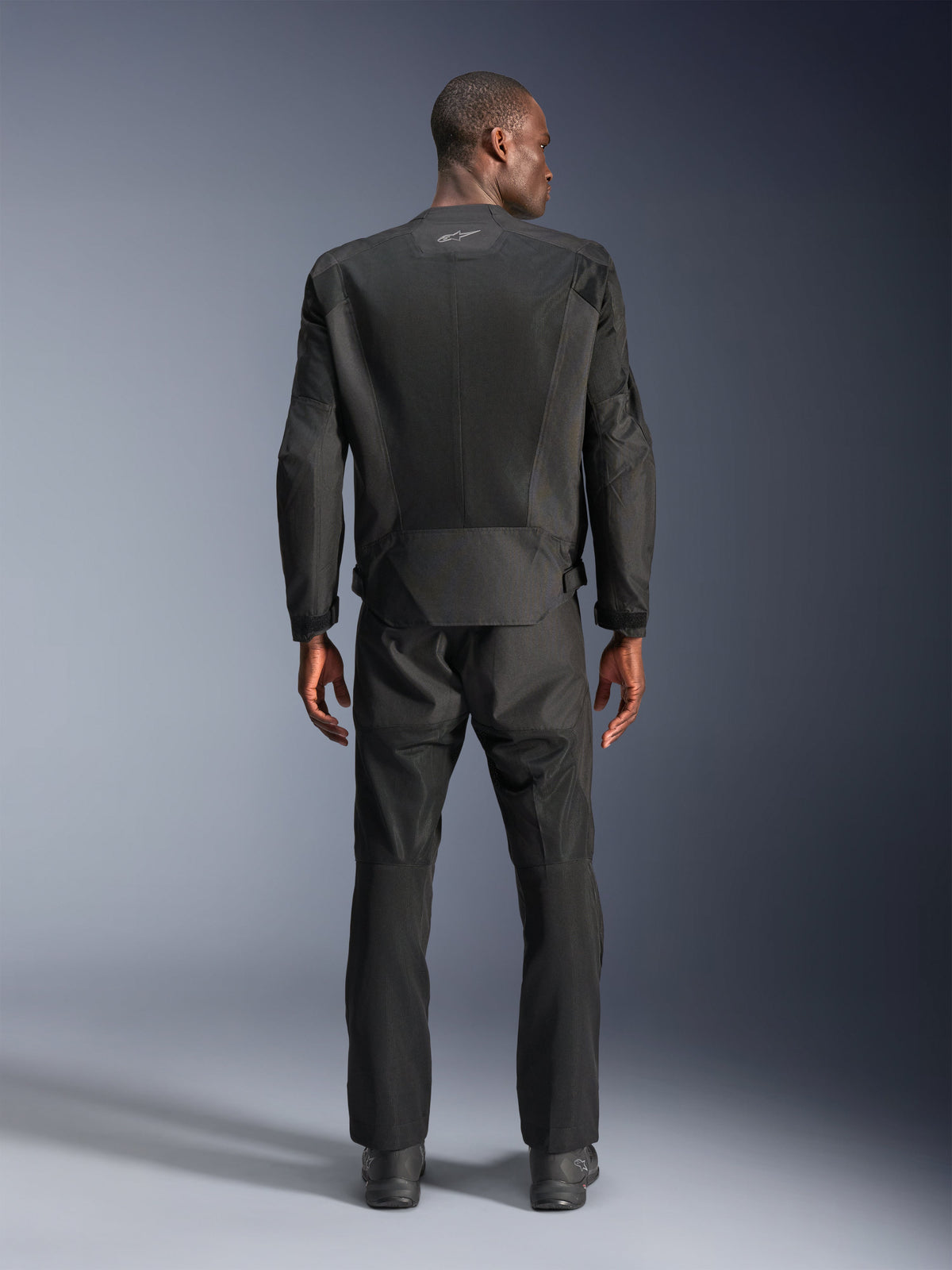 Vue arrière d'un homme portant la veste Alpinestars C-1 Air en noir, veste de moto urbaine avec empiècements en maille technique, pantalon textile et bottes noirs assortis, esthétique épurée avec logo réfléchissant sur le haut du dos.