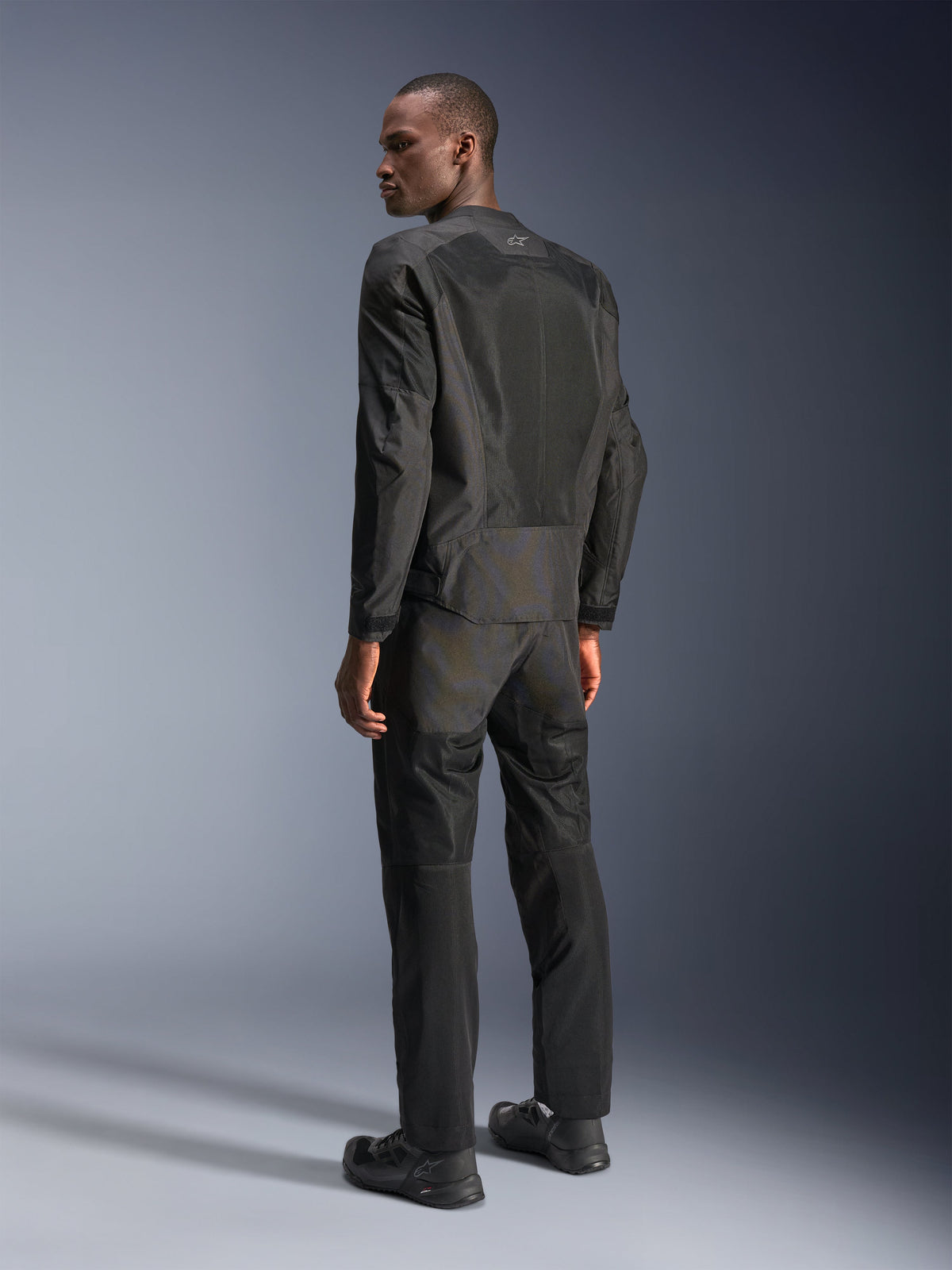 Veste Alpinestars C-1 Air, veste de moto urbaine, noire, vue arrière montrant les panneaux en maille technique pour la circulation de l'air et le logo réfléchissant, portée avec un pantalon et des chaussures de moto noirs assortis, esthétique épurée et raffinée pour les trajets en ville