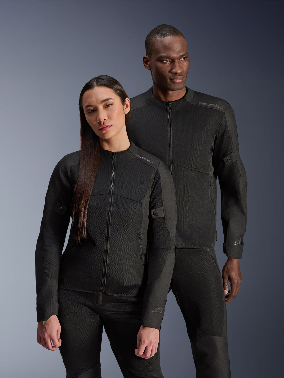 Un homme et une femme portant les vestes Alpinestars C-1 Air et Stella C-1 Air, vestes de moto urbaines assorties en textile noir avec panneaux en mesh pour la ventilation, associées à des pantalons de moto noirs pour un look contemporain et épuré.