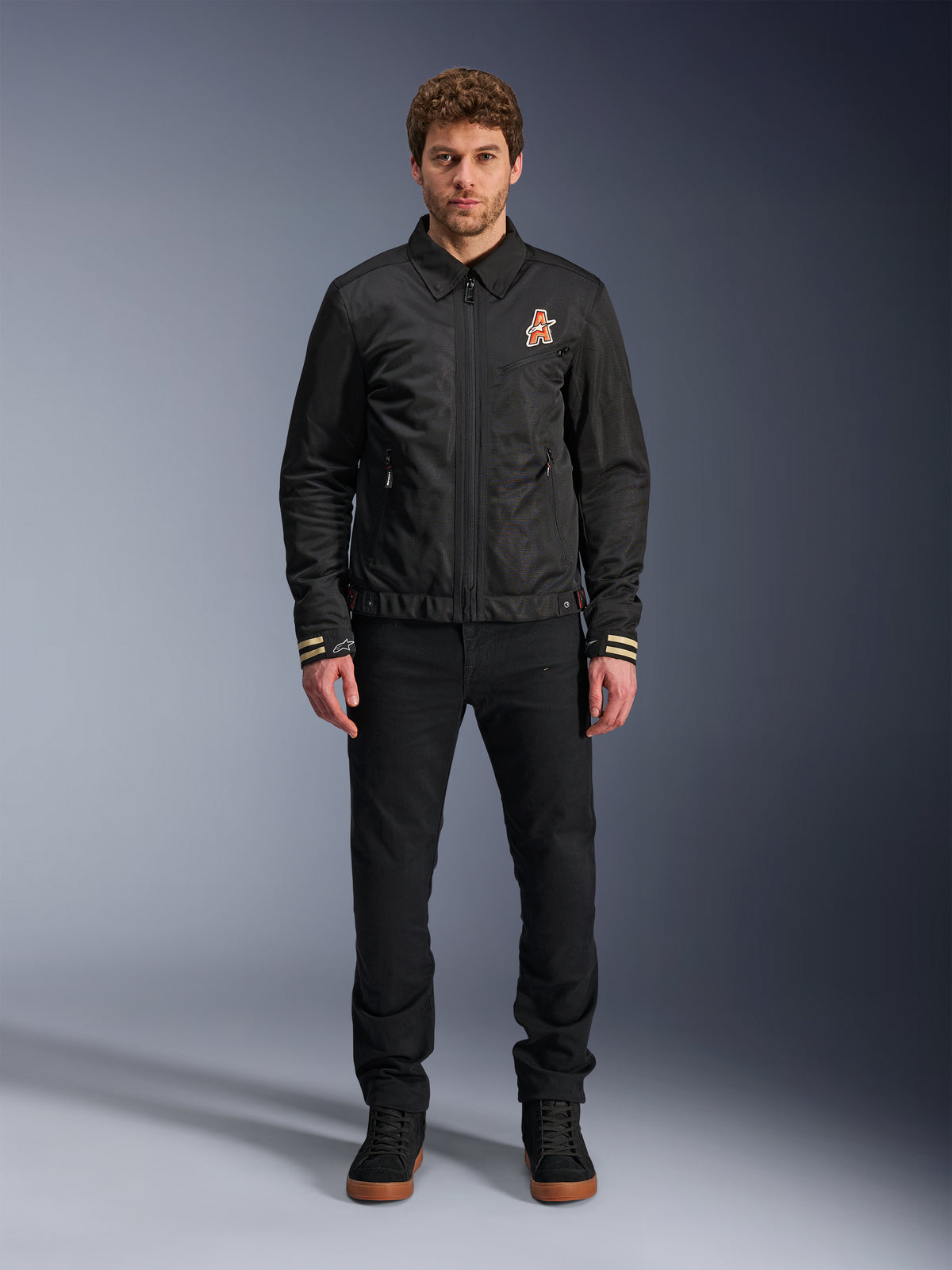 Mannequin homme portant la veste Alpinestars Ignite Air en noir, dotée d'un col chemise classique, d'une fermeture éclair frontale intégrale et d'un logo orange sur la poitrine. Portée avec un pantalon noir et des baskets montantes noires pour un look de moto urbain contemporain.