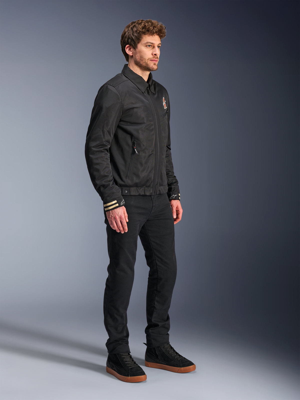 Veste Alpinestars Ignite Air, veste de moto urbaine, noire, portée par un mannequin avec un pantalon noir et des baskets noires, dotée d'un col de chemise traditionnel, de graphismes d'inspiration club sur la poitrine et les poignets, design contemporain élégant avec une finition en maille et satin