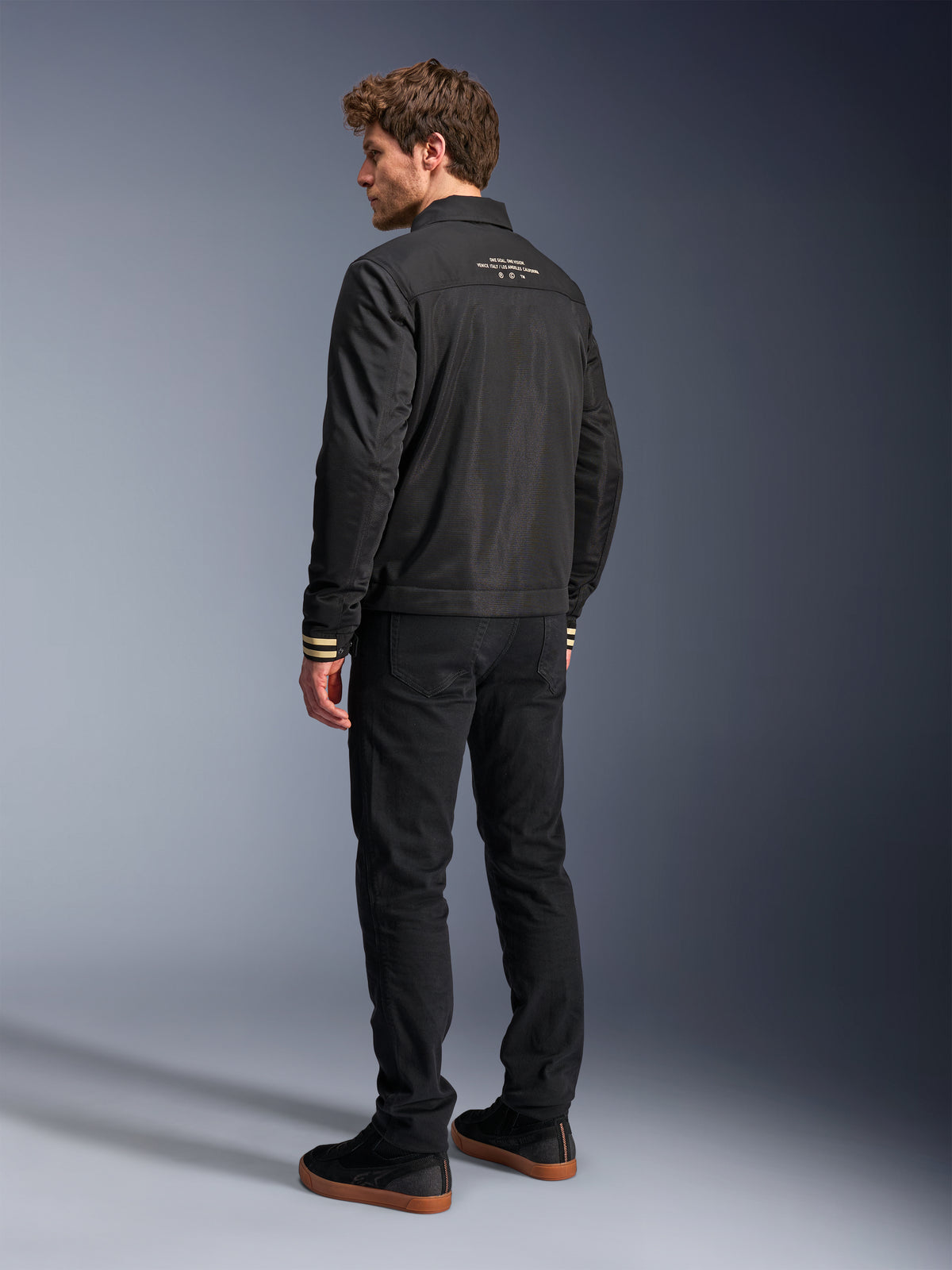 Veste Alpinestars Ignite Air, veste de moto noire, vue arrière présentant un texte graphique blanc sur le haut du dos et des poignets à rayures beiges, silhouette de style Harrington avec corps en maille, associée à un pantalon noir et des baskets noires