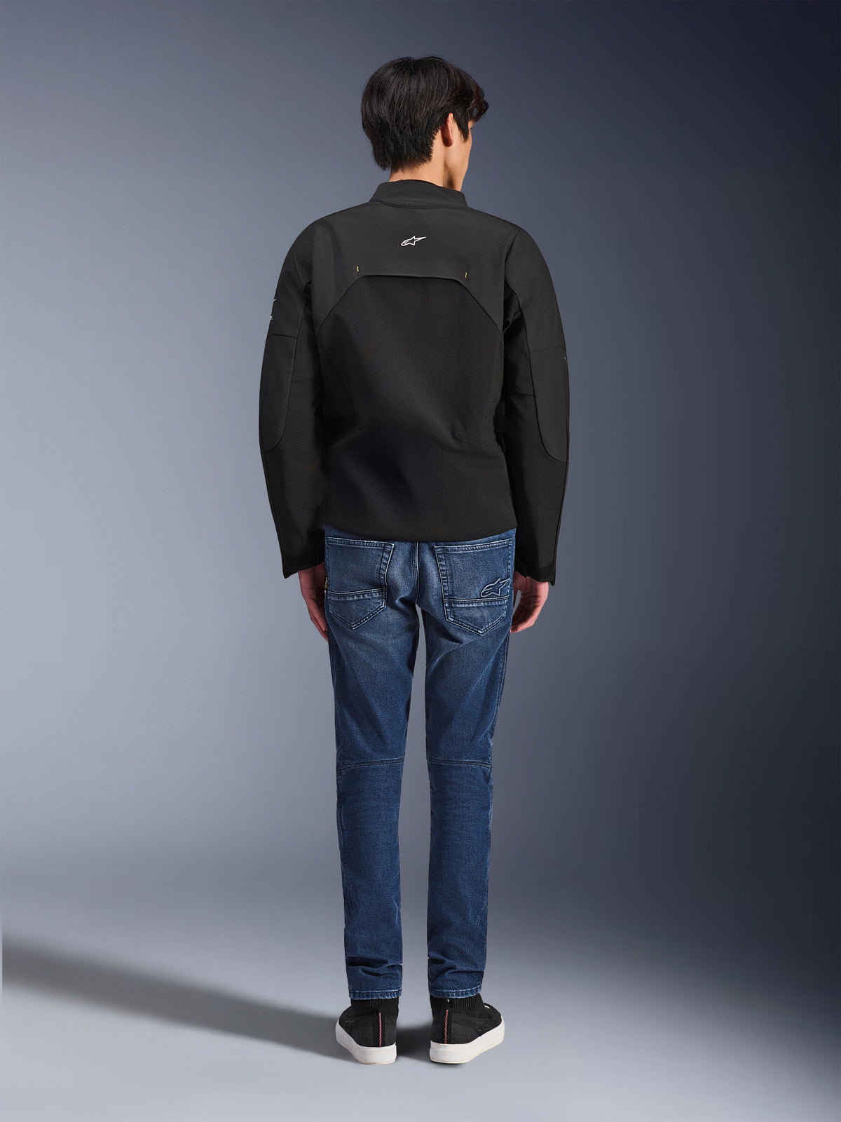 Vue arrière d'un mannequin portant la veste Alpinestars Provoke, veste de moto en textile technique noir, associée à un jean de moto en denim bleu et des baskets noires, montrant la construction du dos et un petit logo Alpinestars blanc, design urbain contemporain