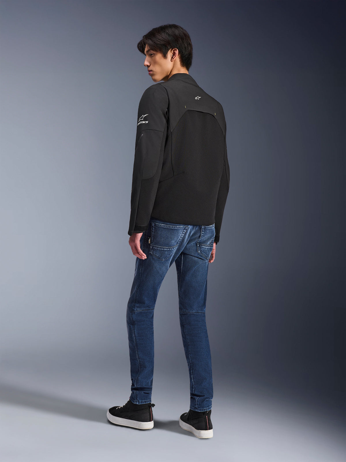 Veste Alpinestars Provoke, veste de moto urbaine noire portée par un mannequin de trois-quarts dos, associée à un jean en denim bleu et des baskets noires, avec des inserts en softshell ripstop et des logos Alpinestars blancs sur la manche et le dos.