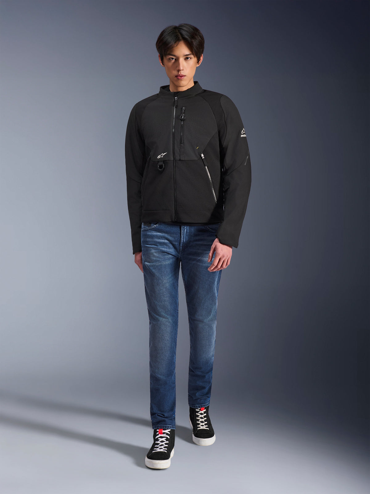 Modèle masculin portant la veste Alpinestars Provoke en noir, veste de moto urbaine en textile dotée de fermetures éclair techniques et d'inserts en softshell ripstop, associée à un jean bleu et des baskets noires, style de motard urbain contemporain.