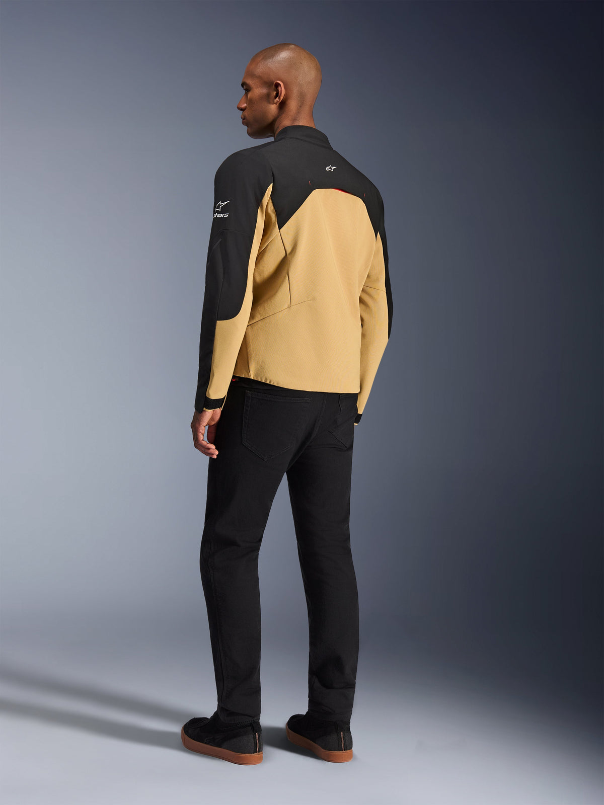 Veste Alpinestars Provoke, veste de moto urbaine en Black Camel, vue de trois-quarts arrière, corps de couleur camel avec empiècements noirs aux épaules et aux manches, logos Alpinestars blancs, portée avec un pantalon noir, équipement de conduite technique contemporain