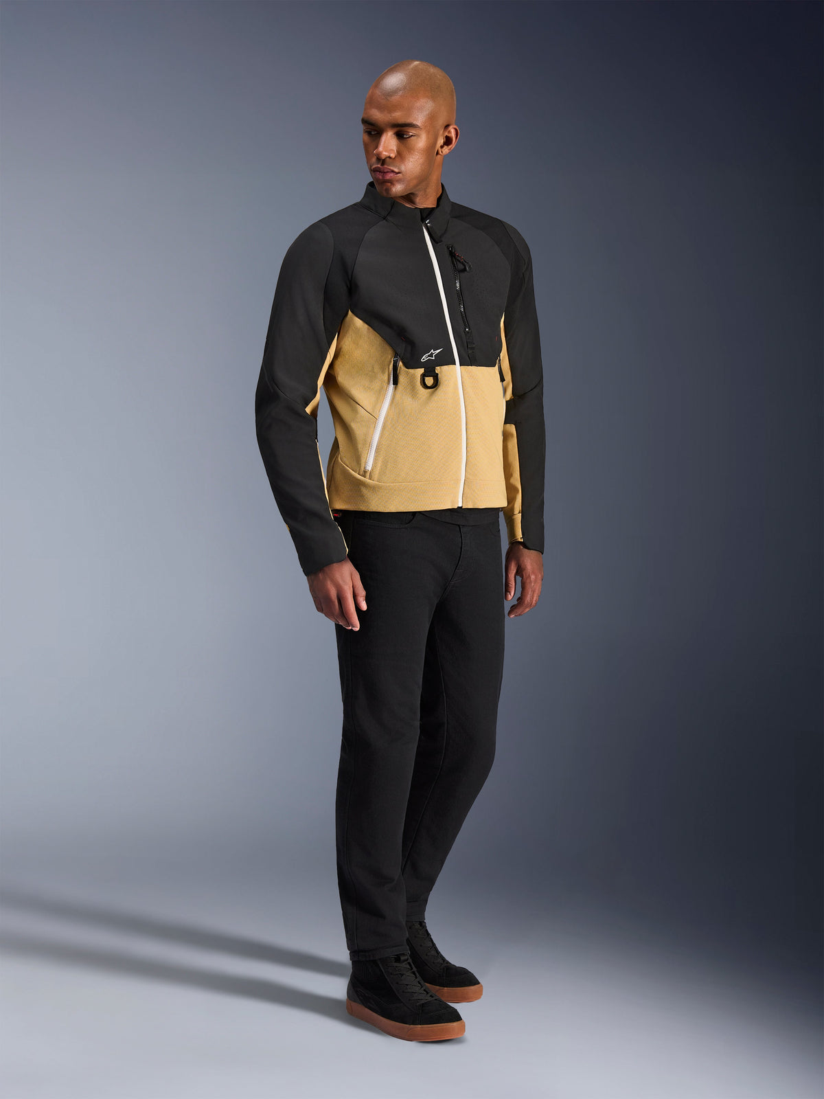 Veste Alpinestars Provoke, veste de moto urbaine, Black Camel, portée par un mannequin avec un pantalon noir et des baskets, design textile bicolore avec le haut noir et le bas du buste camel, détails de fermetures éclair blanches, équipement de moto urbain contemporain